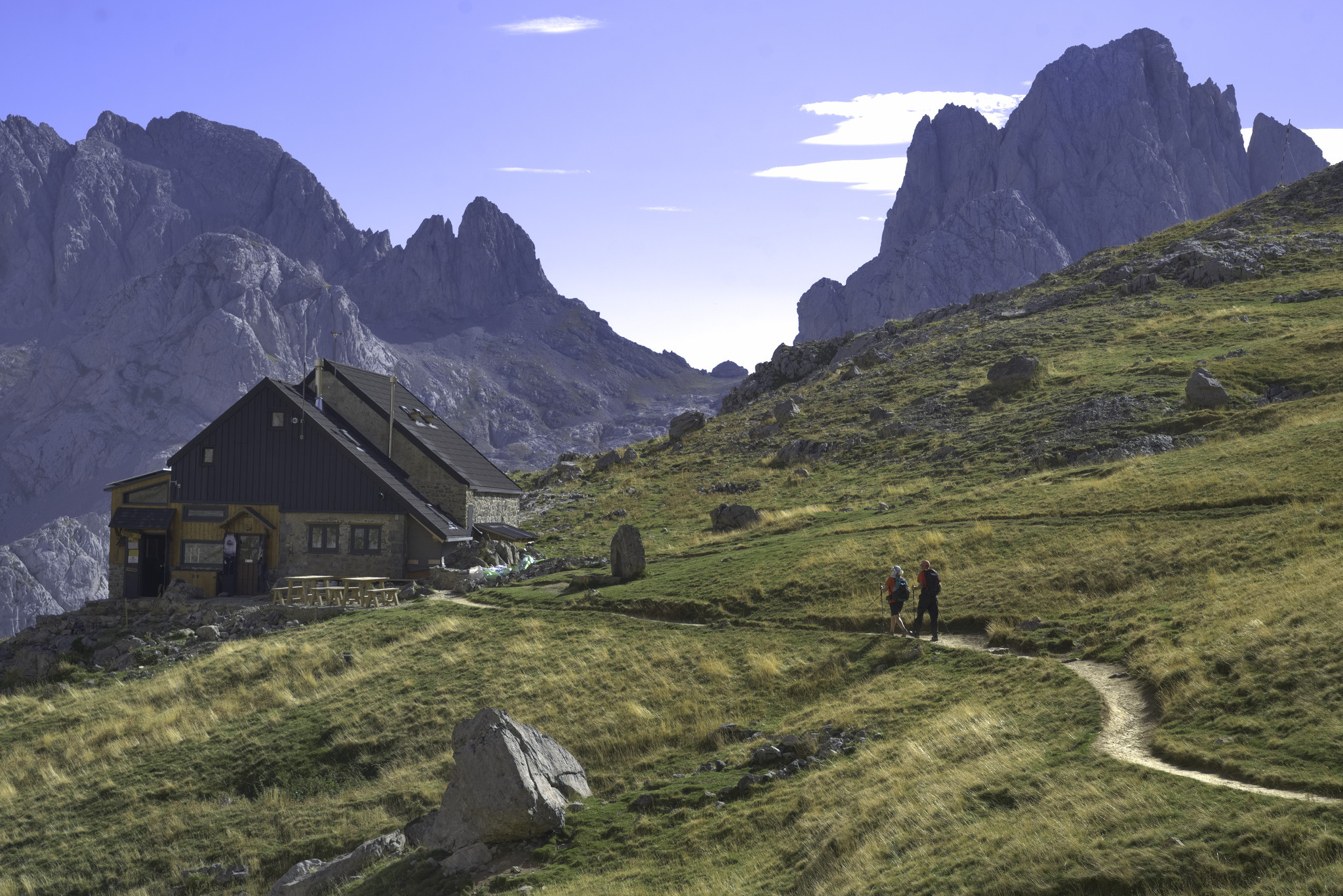 On the left is the Refugio Collado Jermoso, behind are dramatic steep mountains. In the foreground is a grassy expanse with two hikers approaching the hut