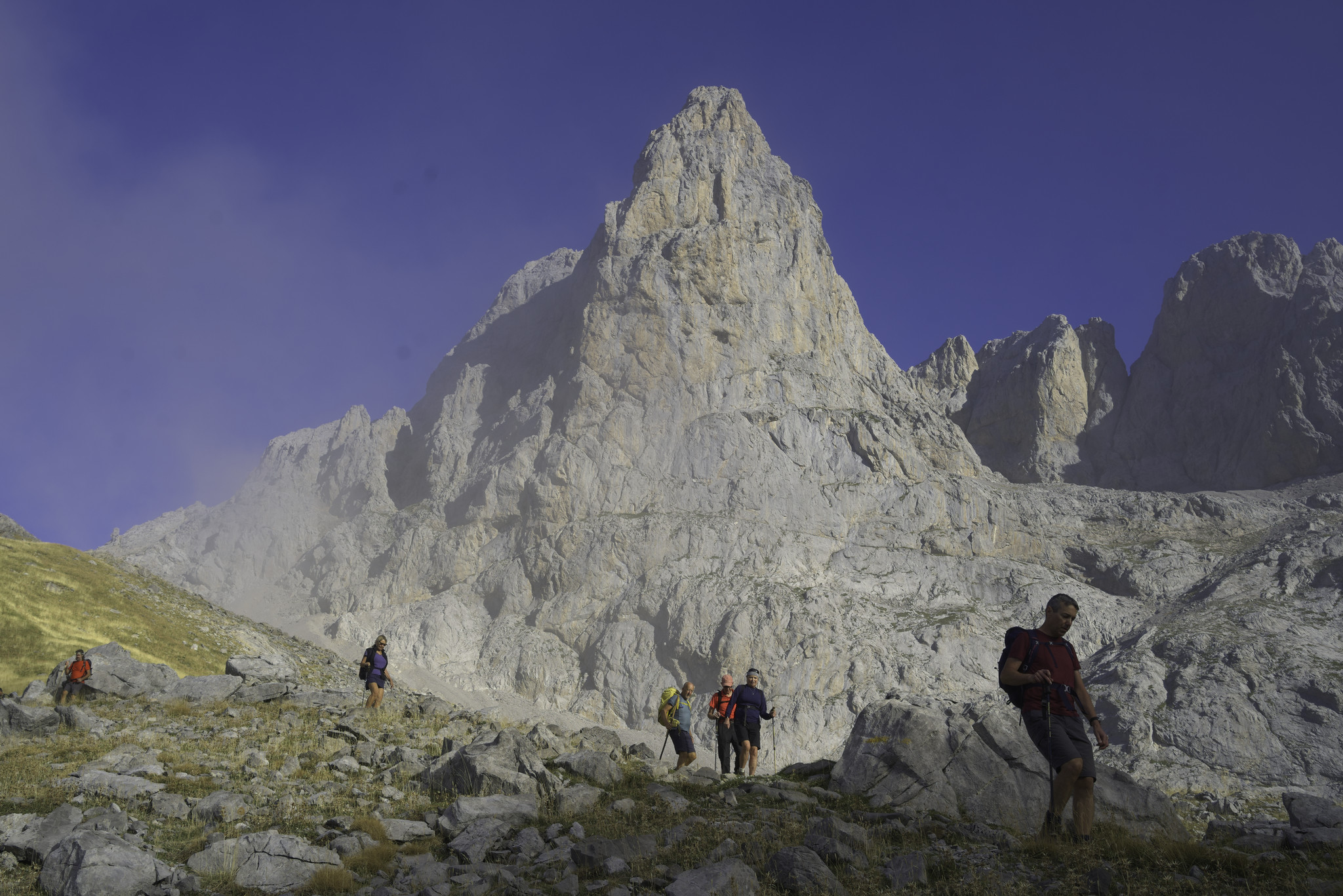 Hikers pass in front of a triangular shaped mountain with a sharp point.