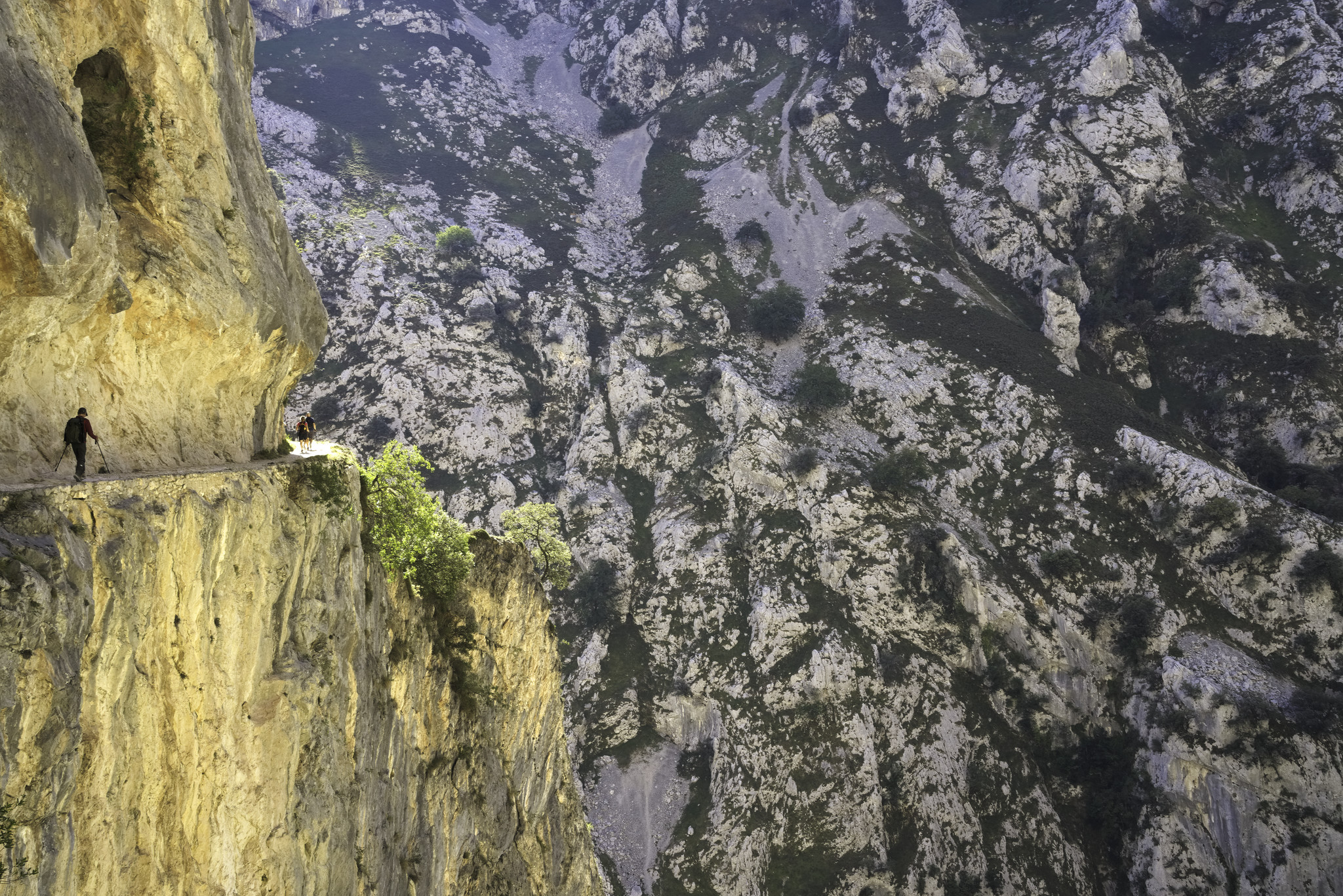 A couple of hikers are seen on a narrow path passing along a vertical yellow rock wall on the left. To the right is a deep gorge