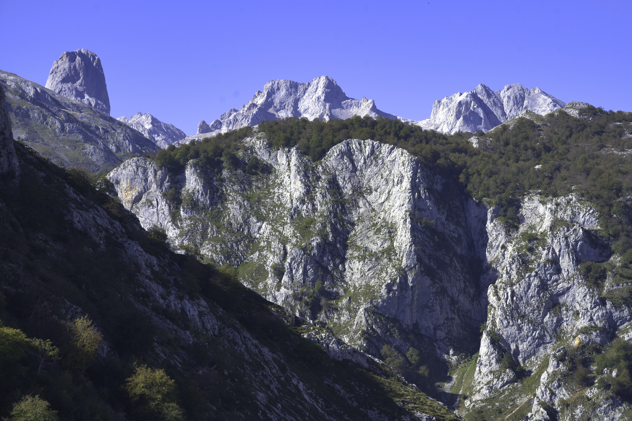 The peak of Naranja de Bulnes dominates the upper left. A range of mountains continues in the background. In the foreground is a sunlit gorge