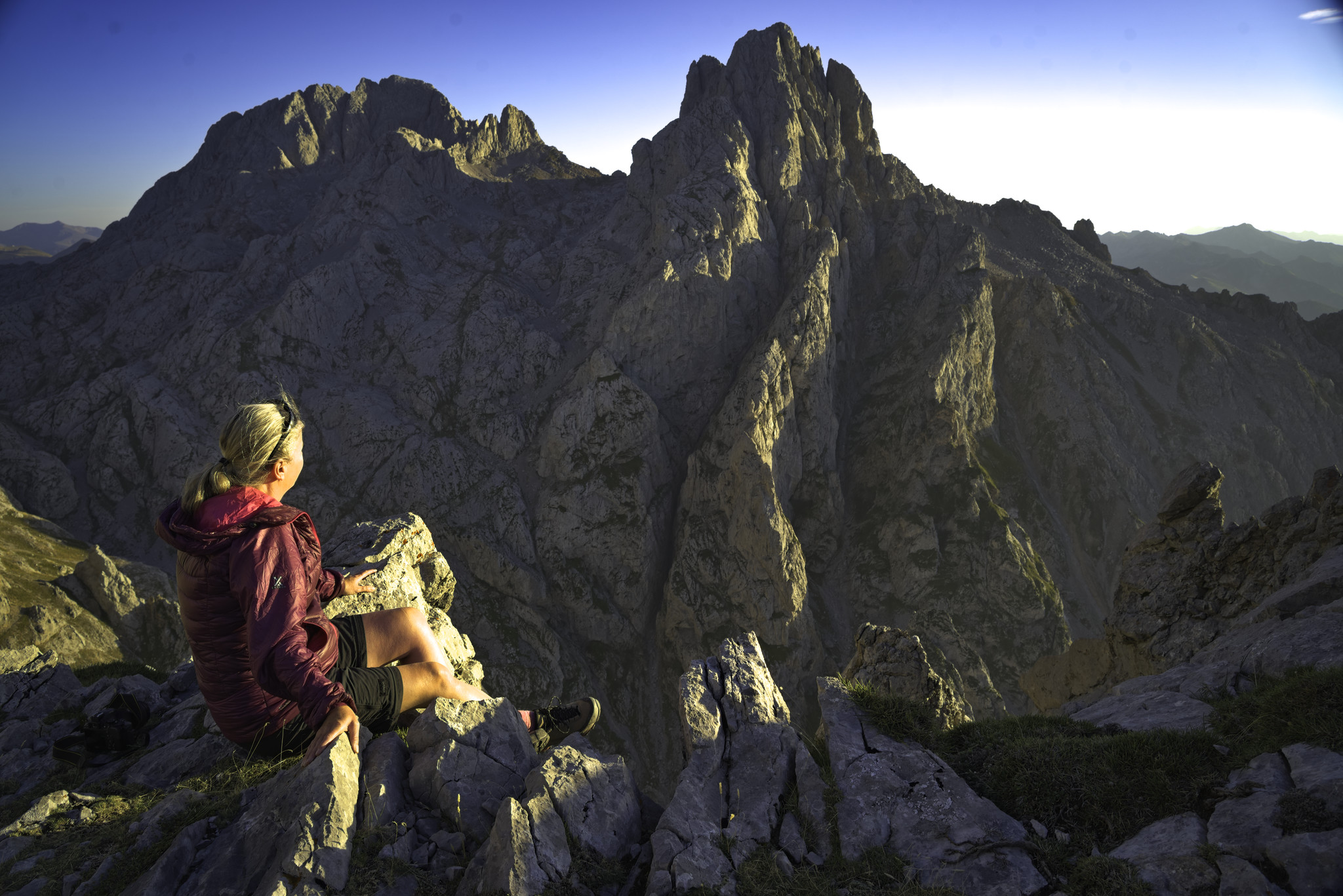 A person with purple jacket is highlighted in the setting sun. behind is a dramatic mountain peak with gorges highlighted in the sun