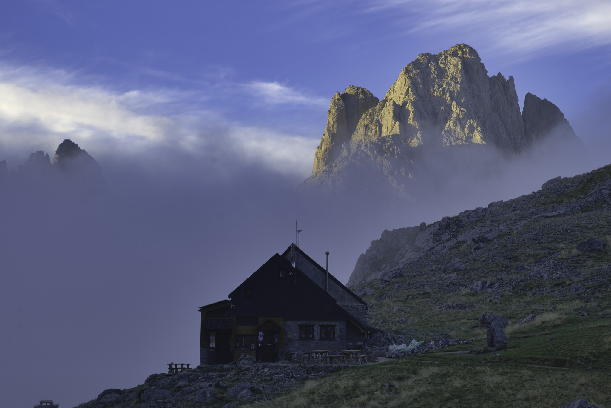 The Refugio Collado Jermoso appears out of the low mountain mist with a huge mountain rising behind it highlighted in the morning sunshine