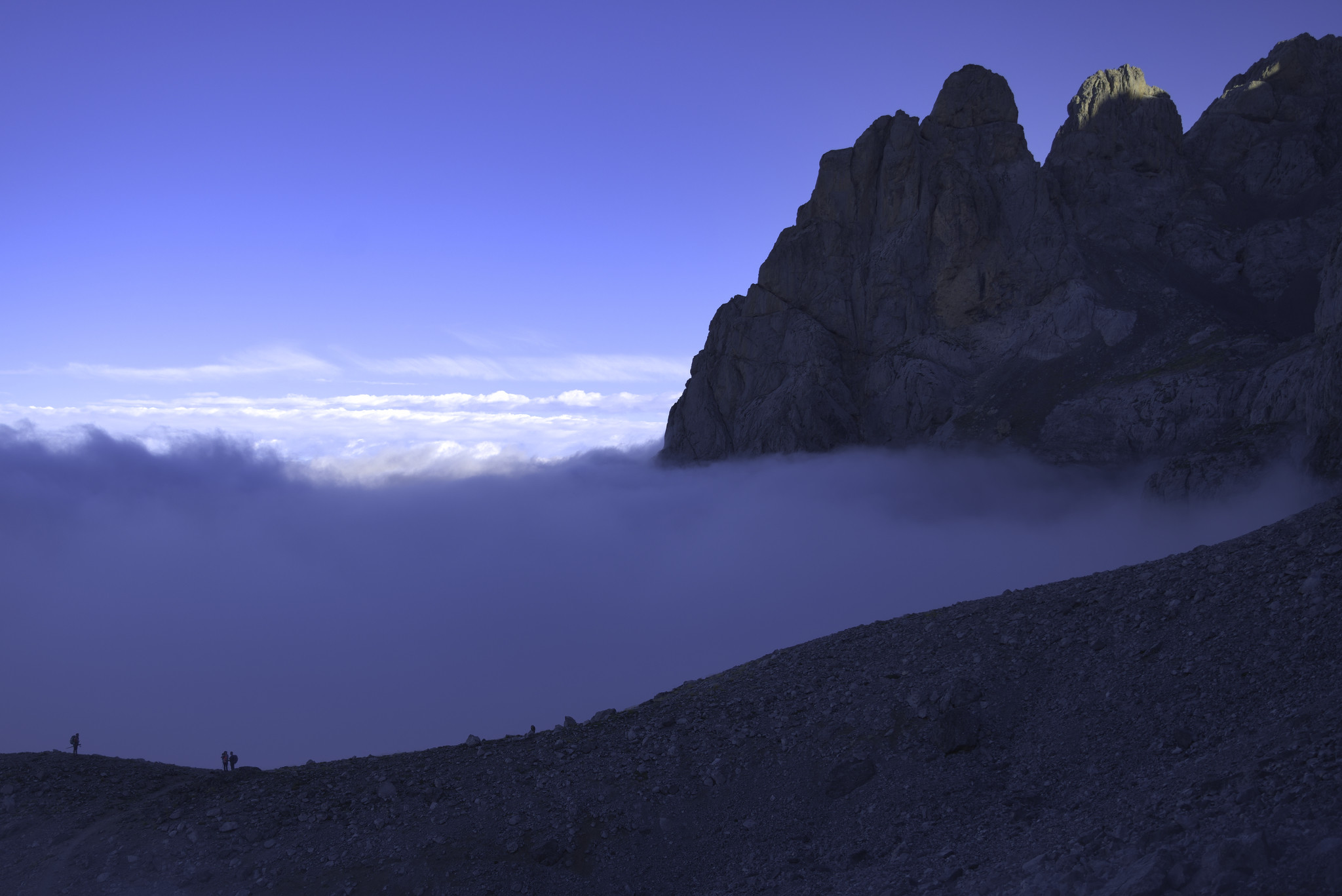 Some hikers are at a small pass on the bottom left of the image. Some low cloud is behind. To the right rises a mountain peak with it's summit highlighted in the morning sun