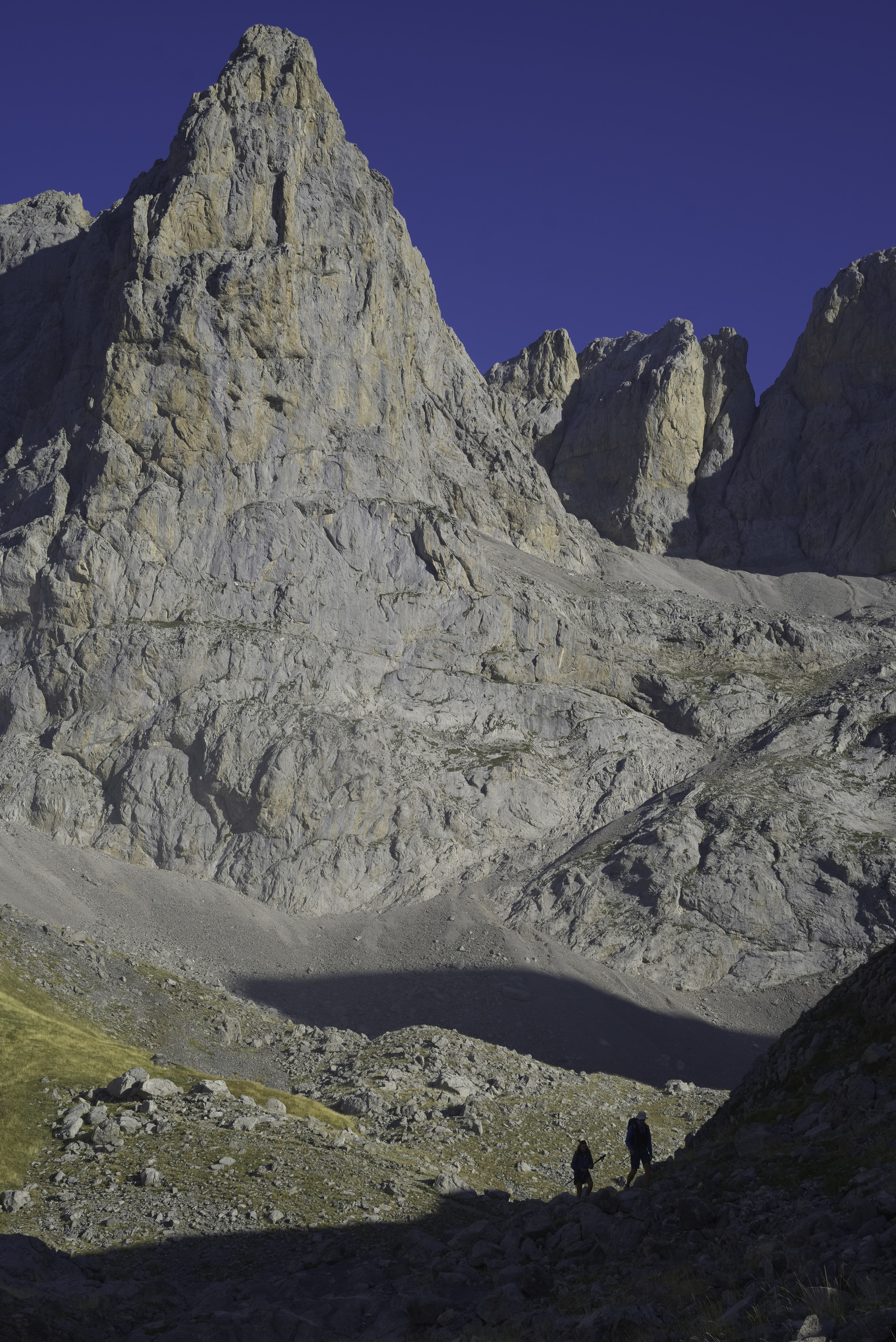The peak of Torre de las Minas de Carbón 2483m rises like a huge pyramid into the sky, clad in gray and orange stone. In the foreground the silhouette of two hikers