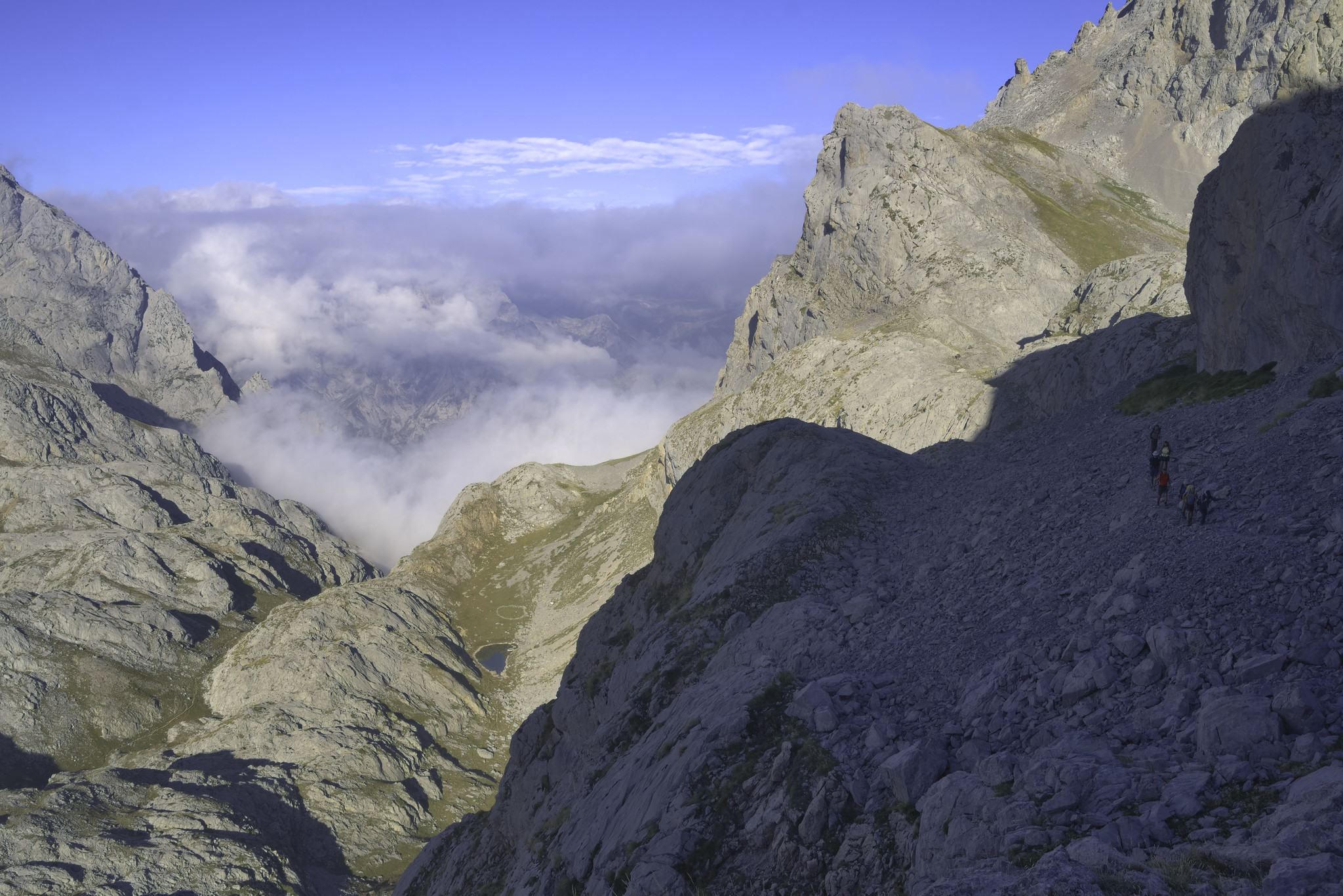 Some clouds gather in the west, blow is a small lake nestled between crags. To the right in shadow are a small group of hikers making their way along a narrow mountain track