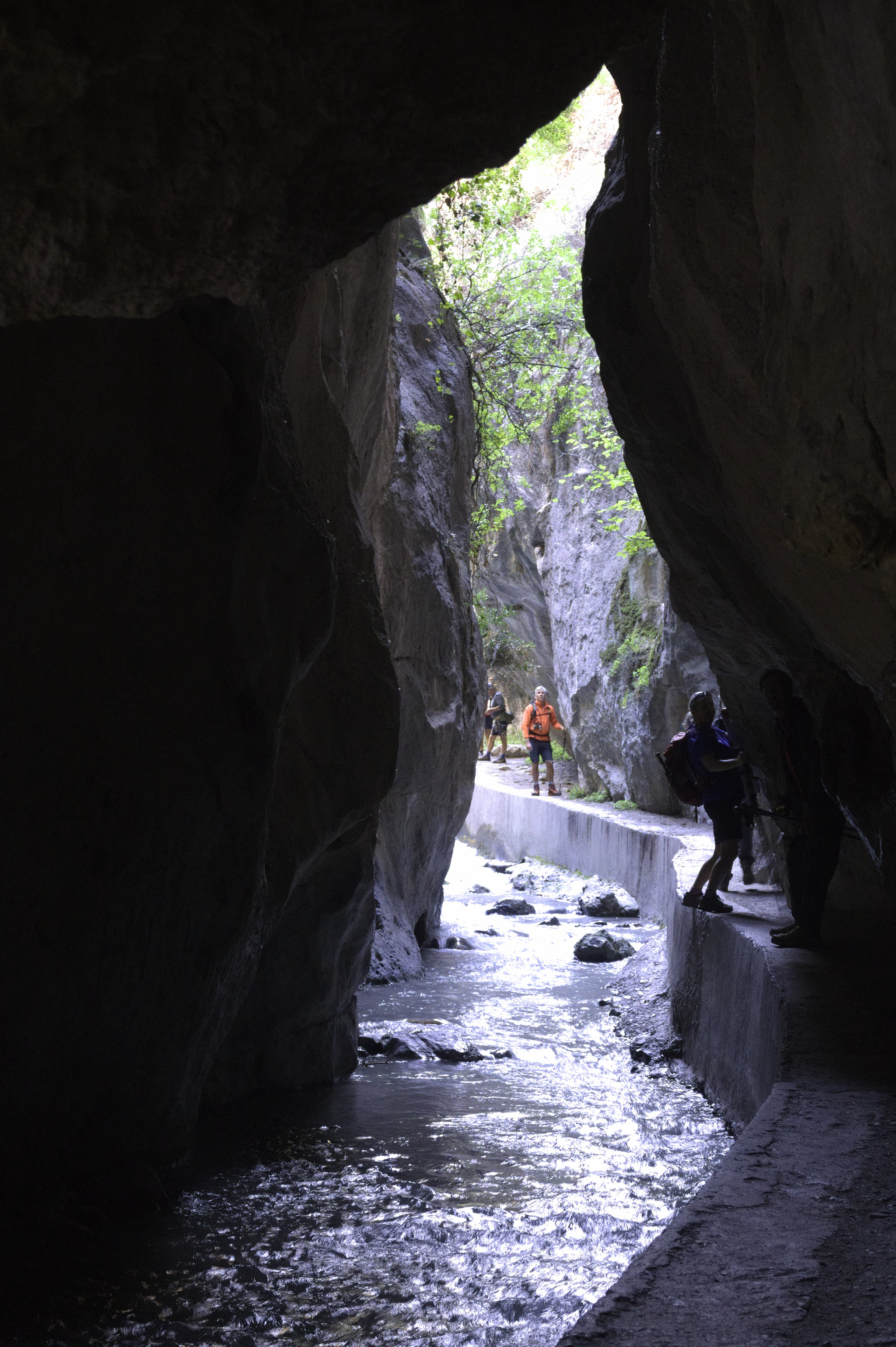 A tunnel in a gorge through which runs a small river. A narrow walkway on the left allows hikers to squeeze through
