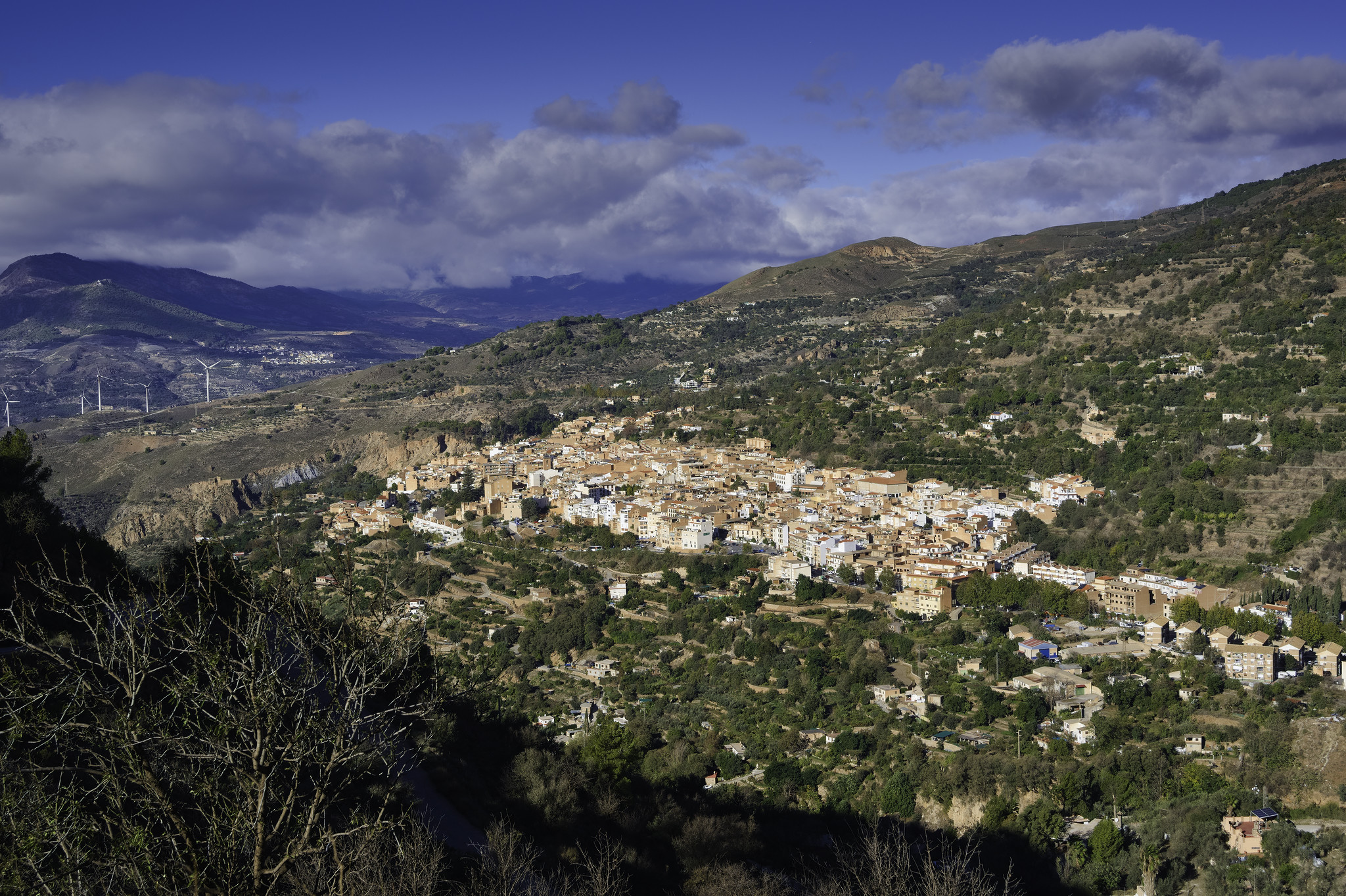 A small town sits in the morning sunshine with clouds behind above some dark hills