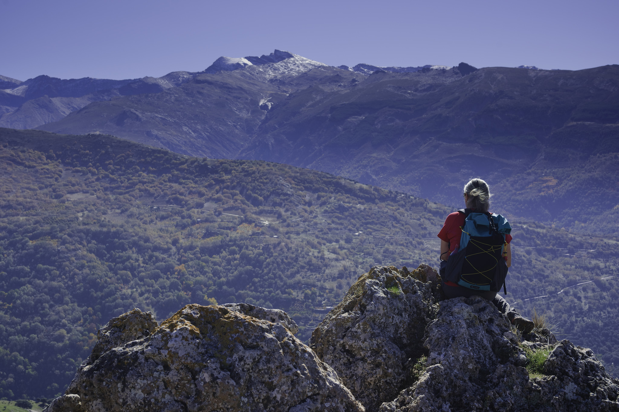 A person sits on a rocky outcrop on the right. In front rise a series of valley reaching up to a snow clad peak