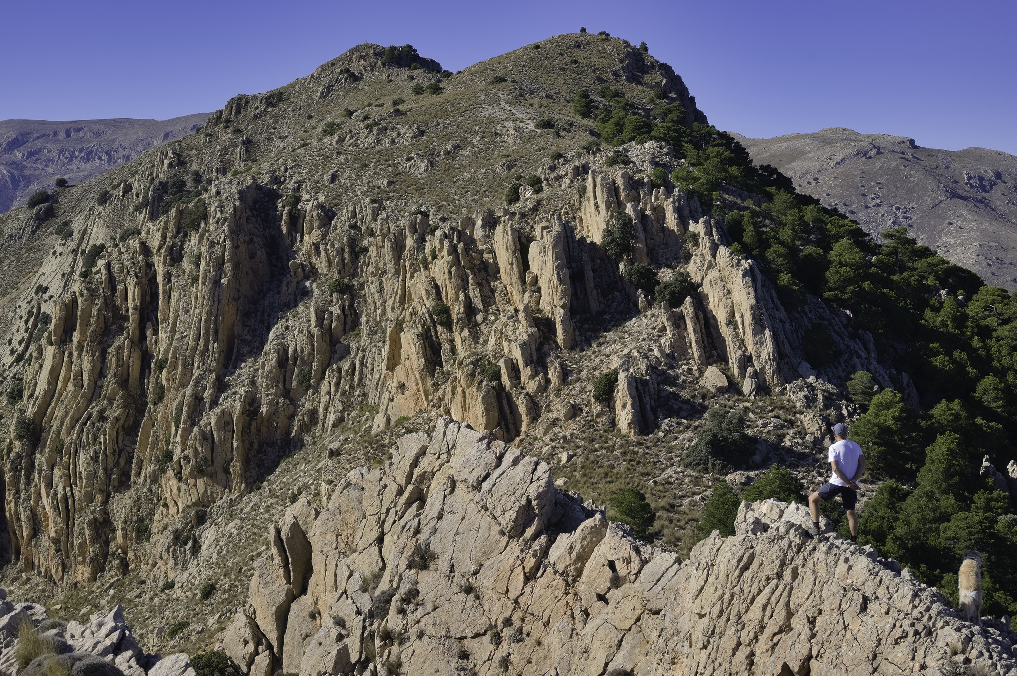 A man in a white t-shirt stands in the bottom left. Behind him is a complicated face of rock walls and fissures leading to a summit with blue sky above.
