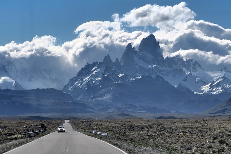 A small car with light on is on a tarmac road that stretches out in a straight line towards some distant mountains. The jagged mountain summits are surrounded by clouds, blue sky above