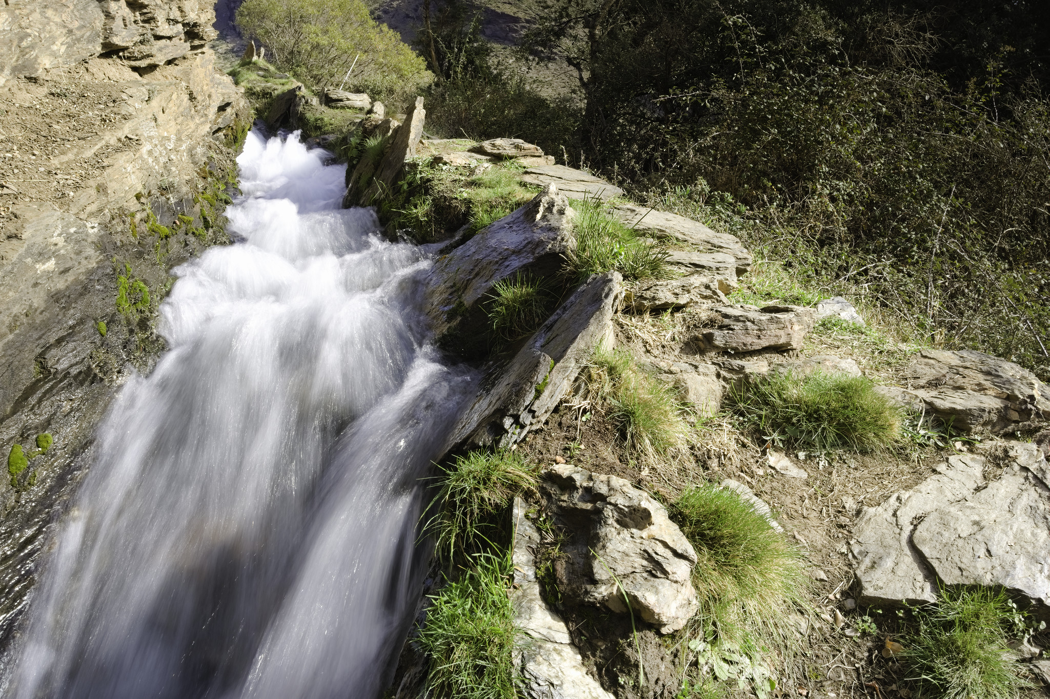 White water gushes down an irrigation channel in the spanish mountains alongside a path
