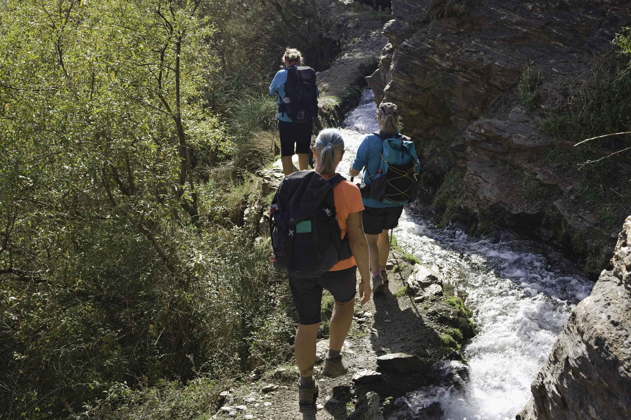 Hikers walk along a narrow trail with an irrigation channel flowing with water alongside. Lots of green shrub and trees to the left, rock walls to the right