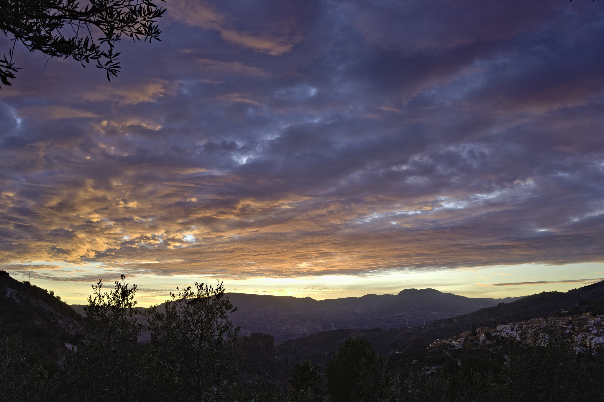 The sky is covered with orange cloud tinged with yellow by the setting sun. Patches of blue sky still show through. Outline of some trees and to the bottom right the lights of a town are seen