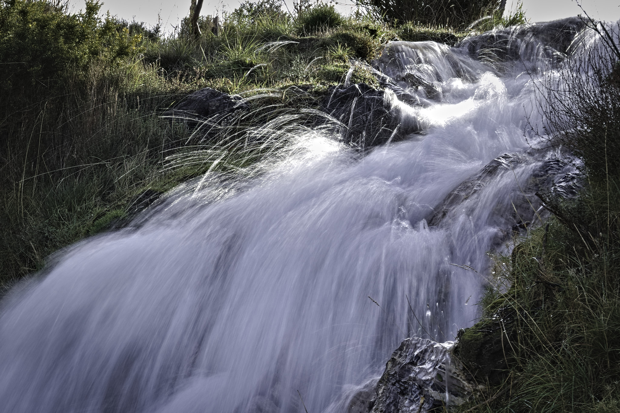 A cascade of water tumbling down a mountain side.The sun illuminates the water at the top. To the left and right are green banks of shrub