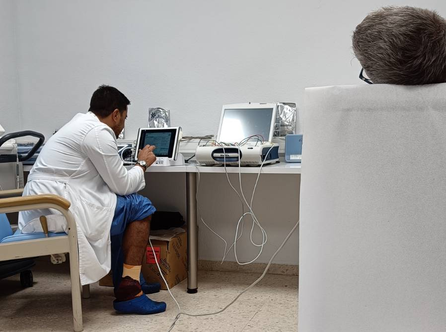 A doctor sits at a table looking at a computer screen while a patient's head is on a bed to the right