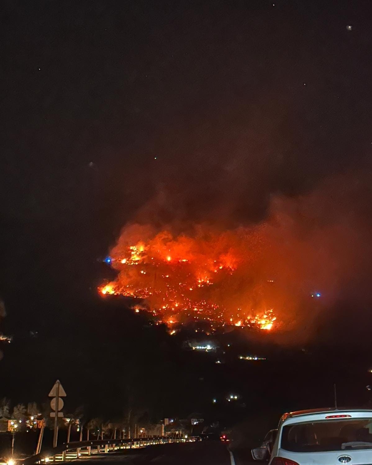 A car passes along the road bottom right. Above, a wildfire burns yellow and orange on the mountainside