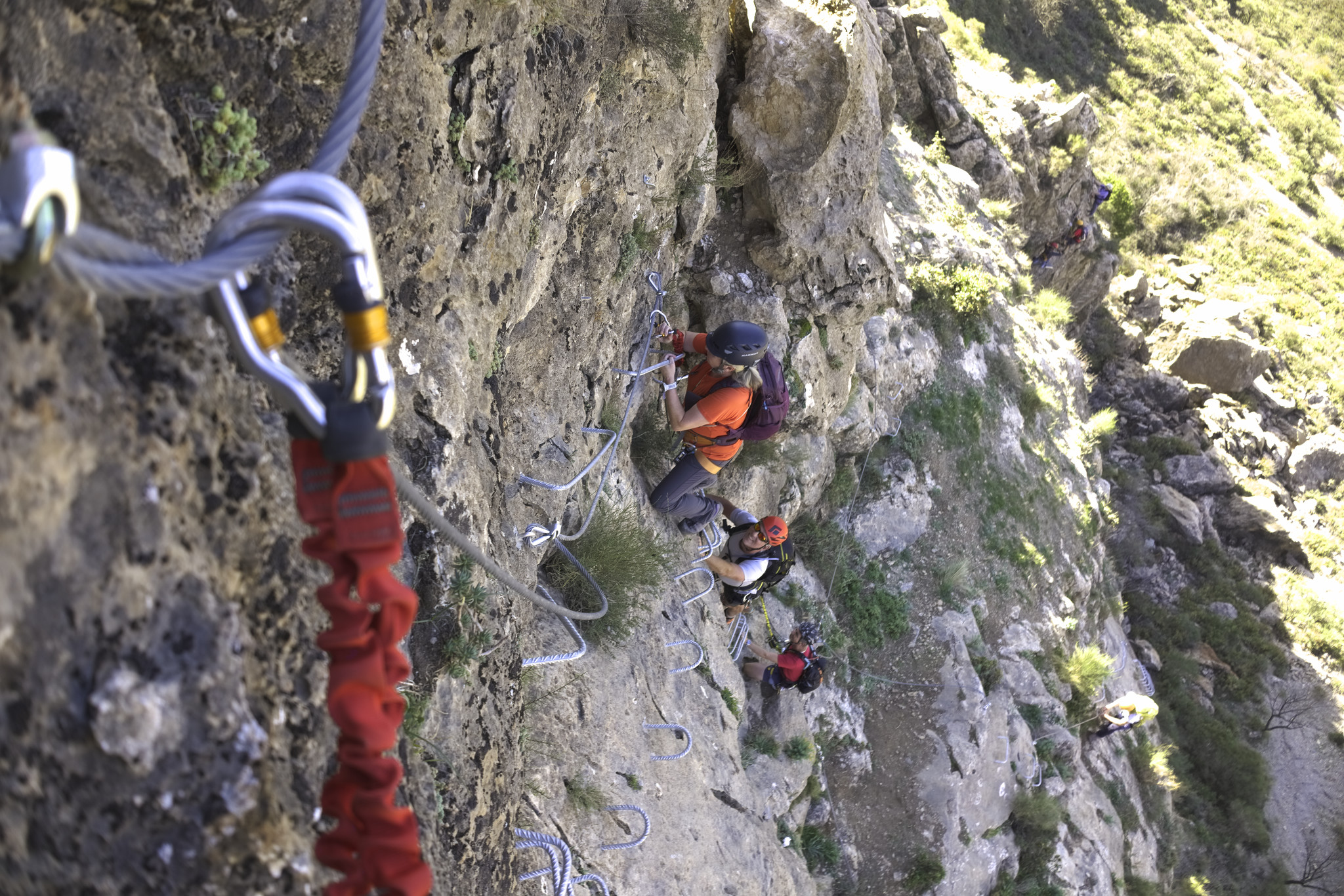 Some people are climbing up or rock wall equipped with ladders and are using cables for protection