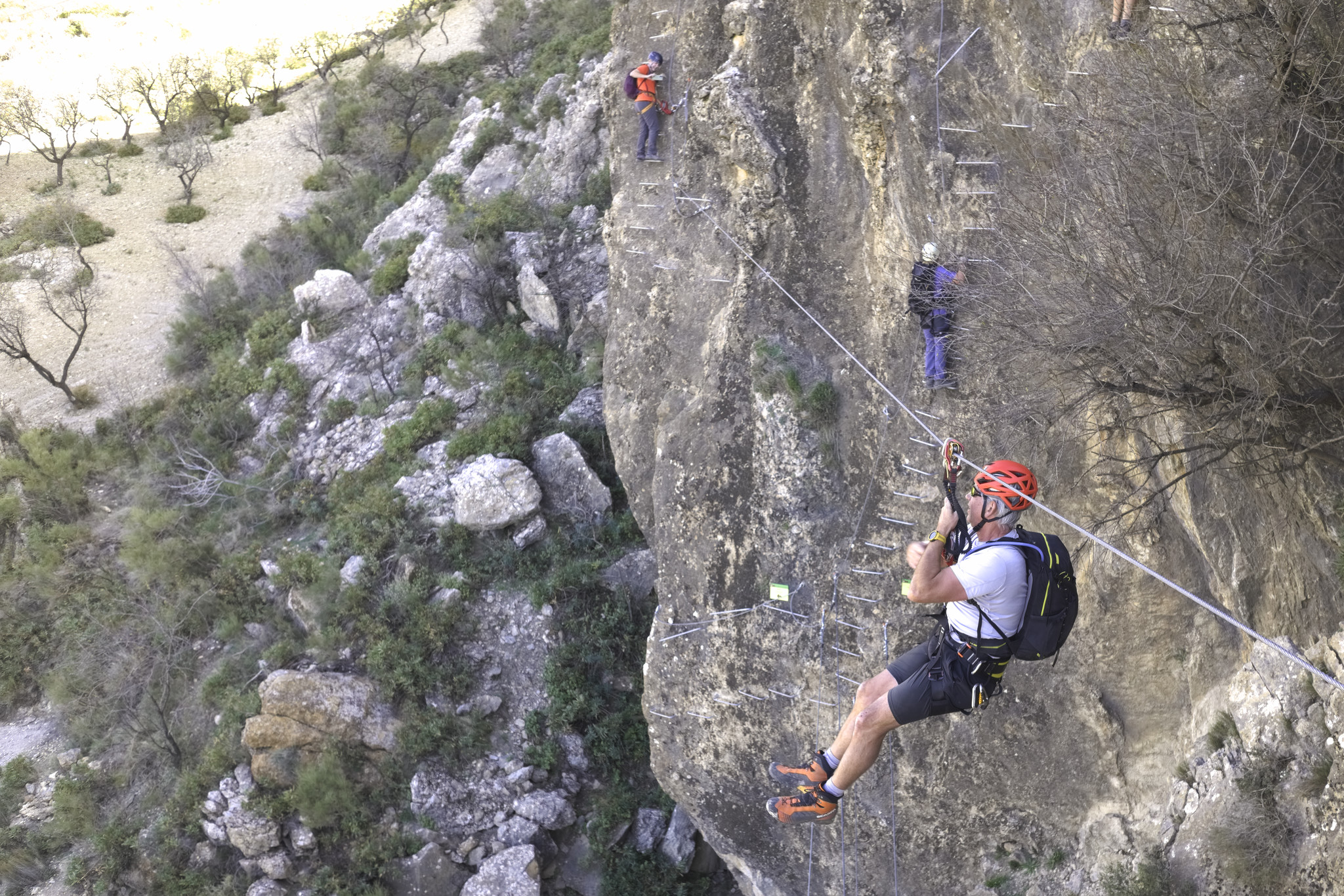 A person swings from a tyrolean traverse zip line whilst others climb steep ladders up the rock wall opposite