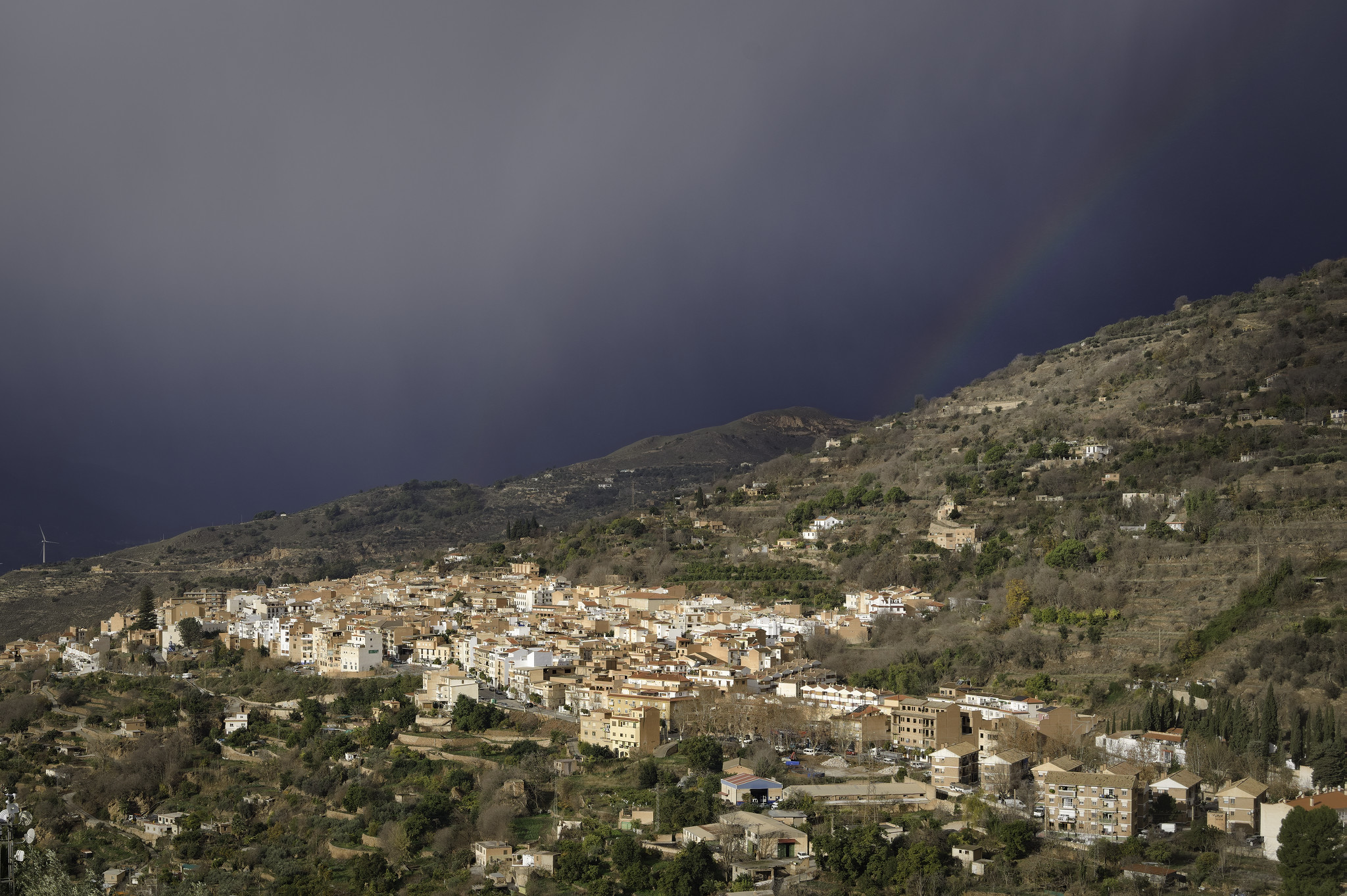 A small town sits in sunlight on a small plateau. Above are dark rain clouds with a small rainbow on the upper right hand side