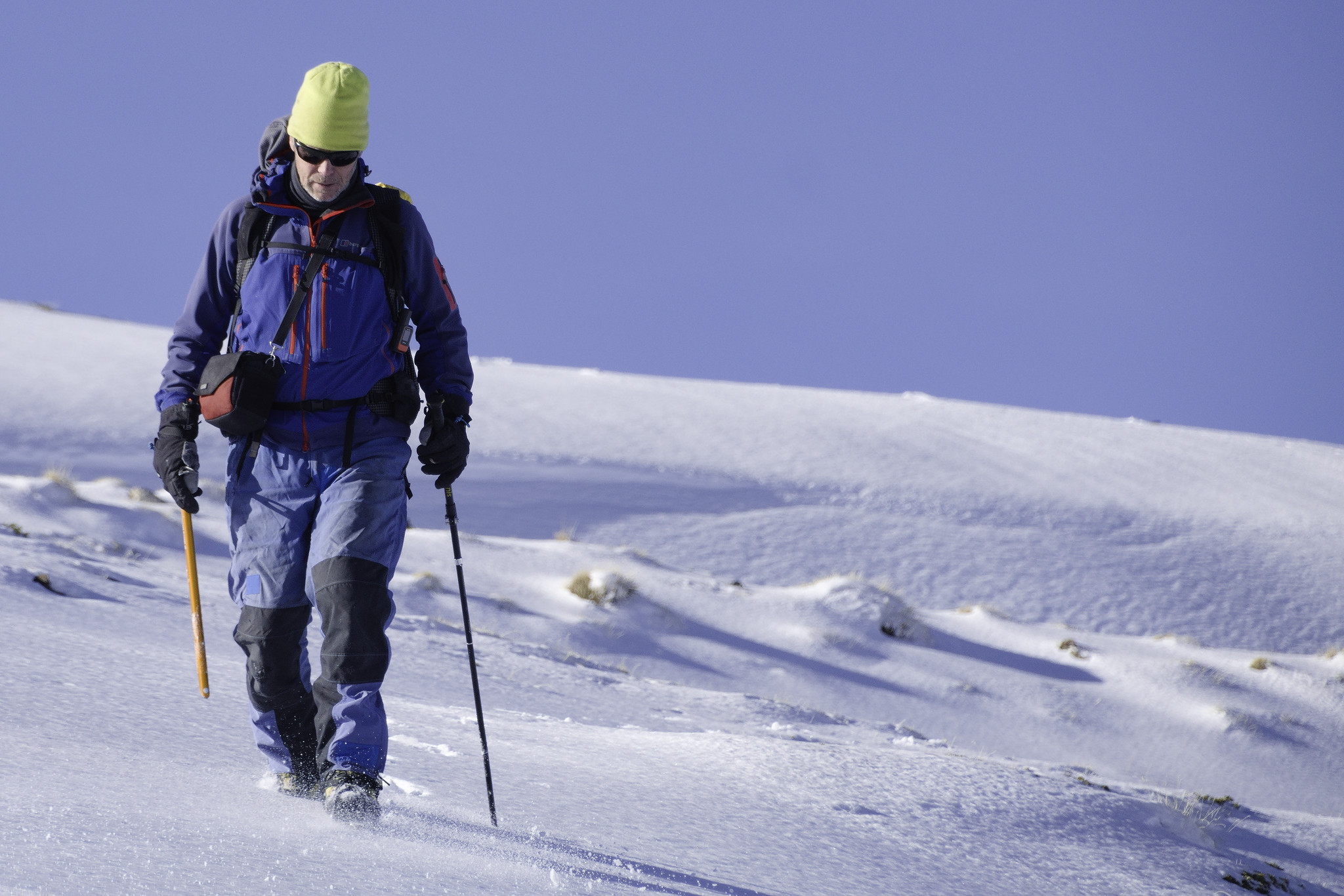 A person (me) with a yellow hat and blue jacket and trousers is walking down a snowfield with an ice axe in one hand and a walking pole in the other.