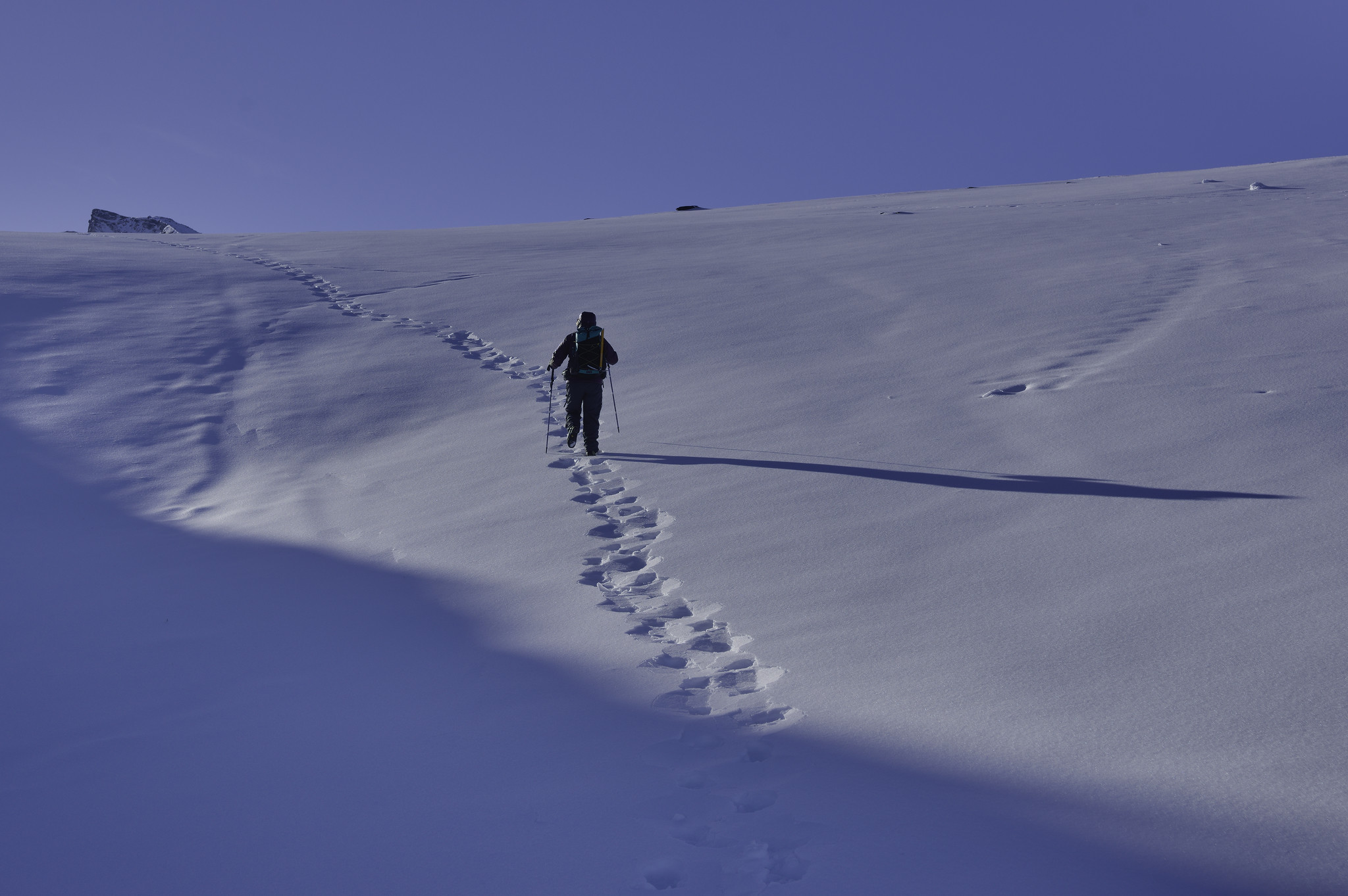 A person follows a solitary pair of tracks up a snow covered hillside. A long shadow comes out of the snow to the right of the person. Above are the indications of mountain summits. Bright blue skies.