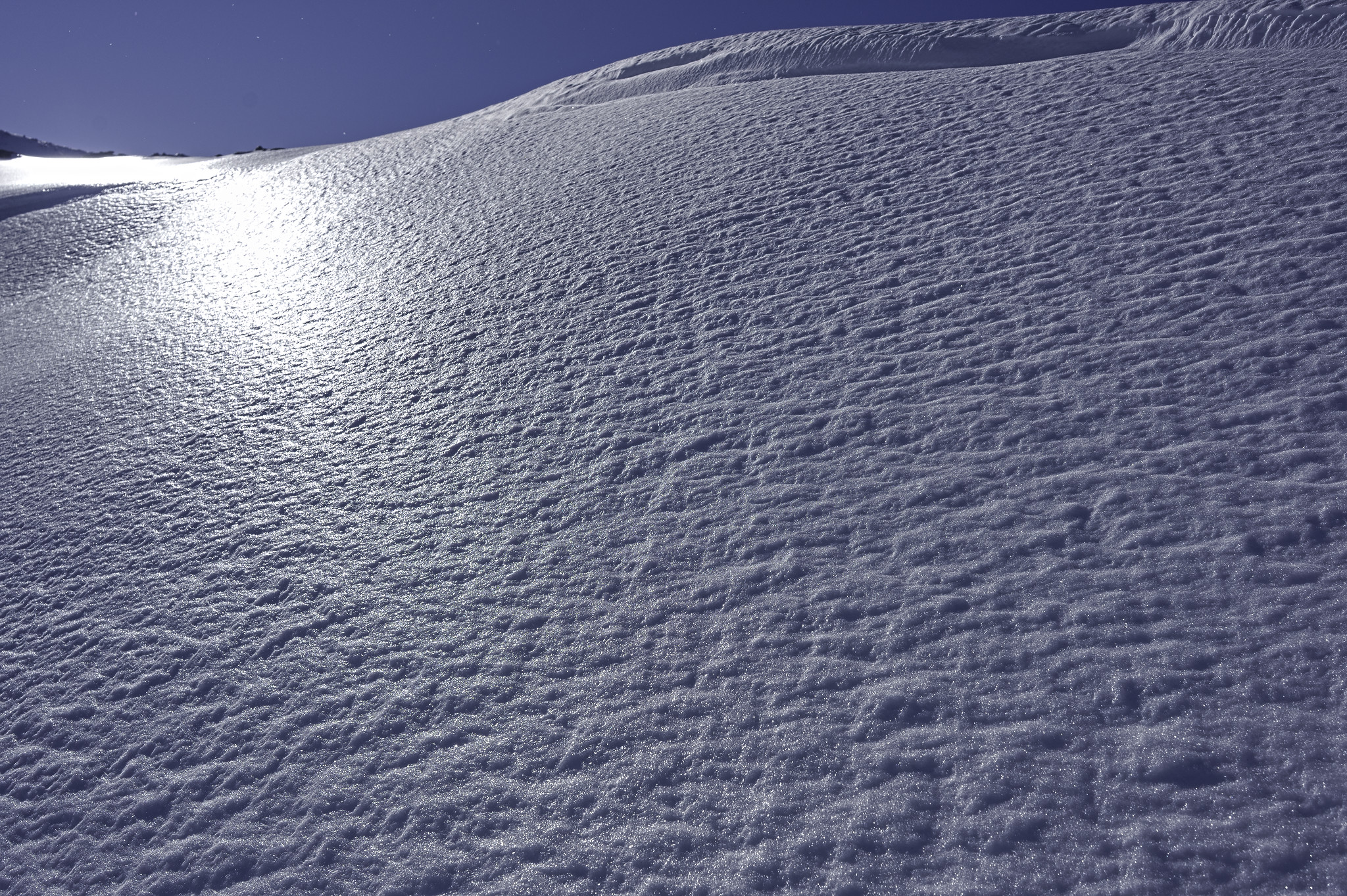 A bare snow slope glints with ice in the early morning sun. Some cornices line the upper right edge of the image