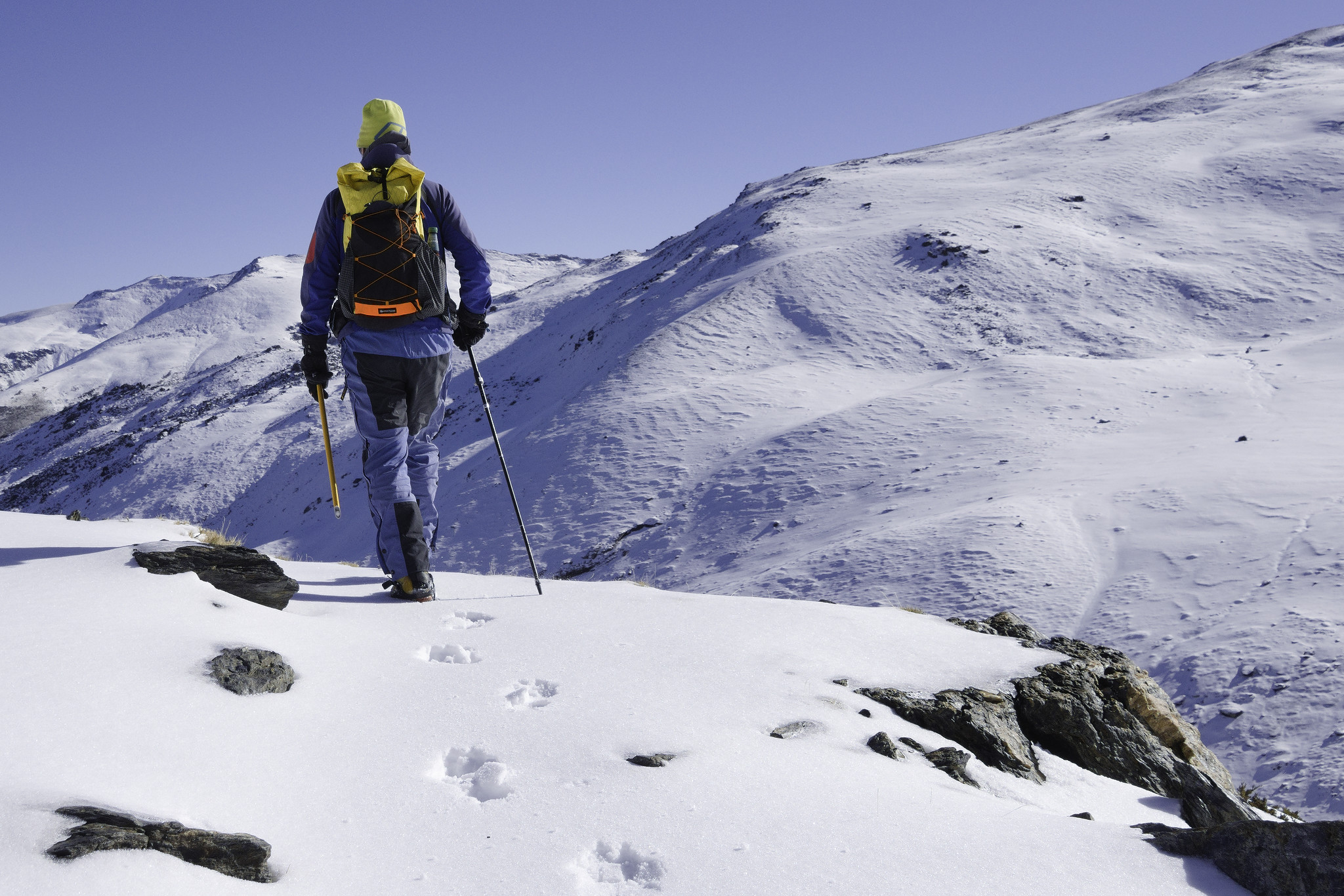 A person in blue with a yellow and black rucksack makes first tracks along a snow covered path. To the right is a valley and then a much larger snow covered mountain rises
