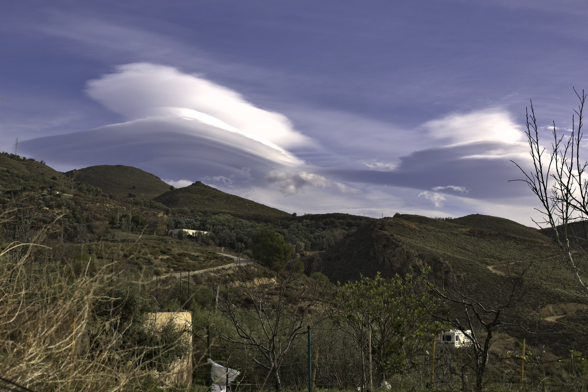 Hills and fields of shrub and farmland are overshadowed by some huge clouds that form into lenticular shapes in the sky