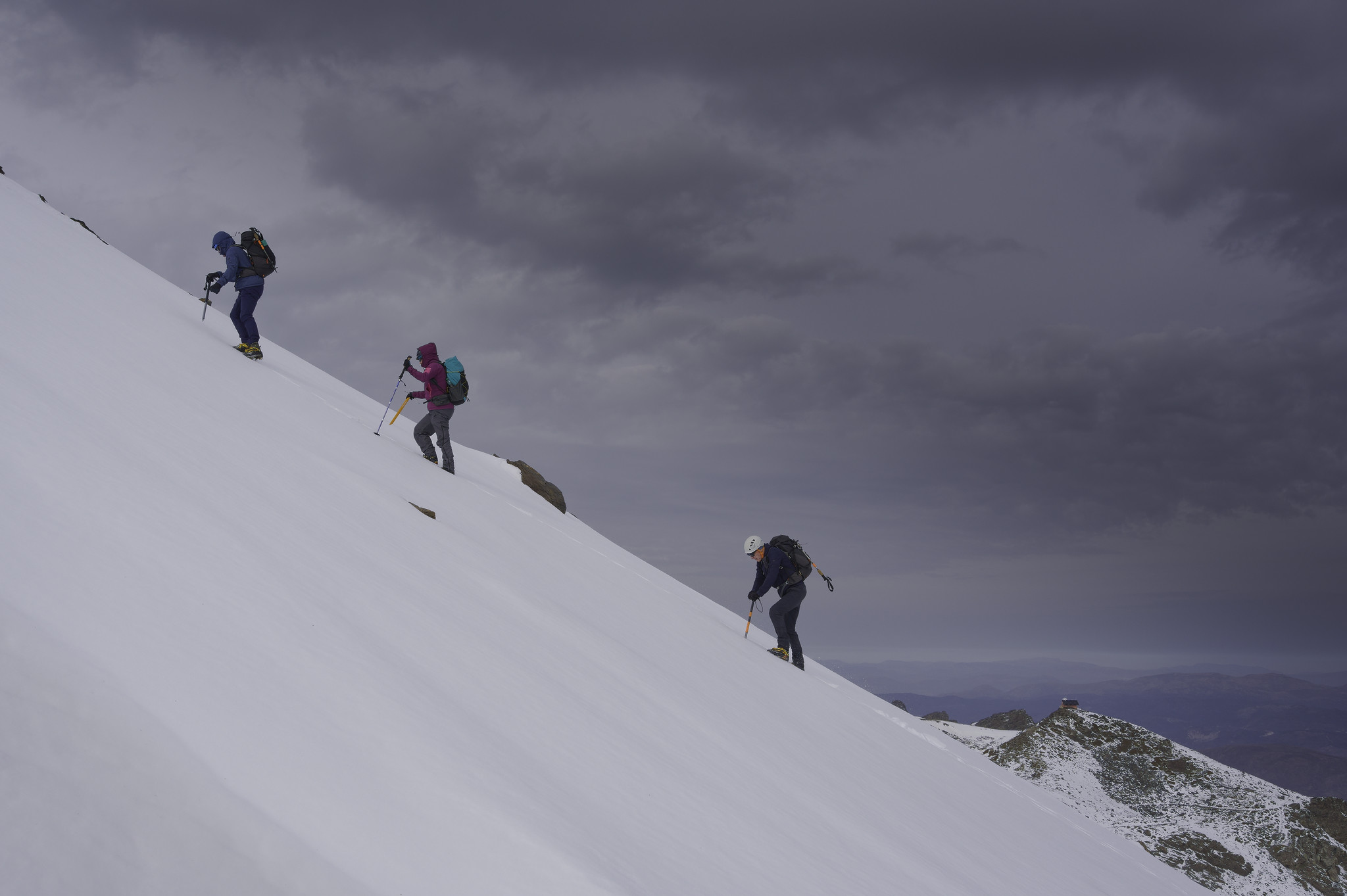 Three people are climbing up a steep snow slope. Behind them dark skies bring the onset of bad weather
