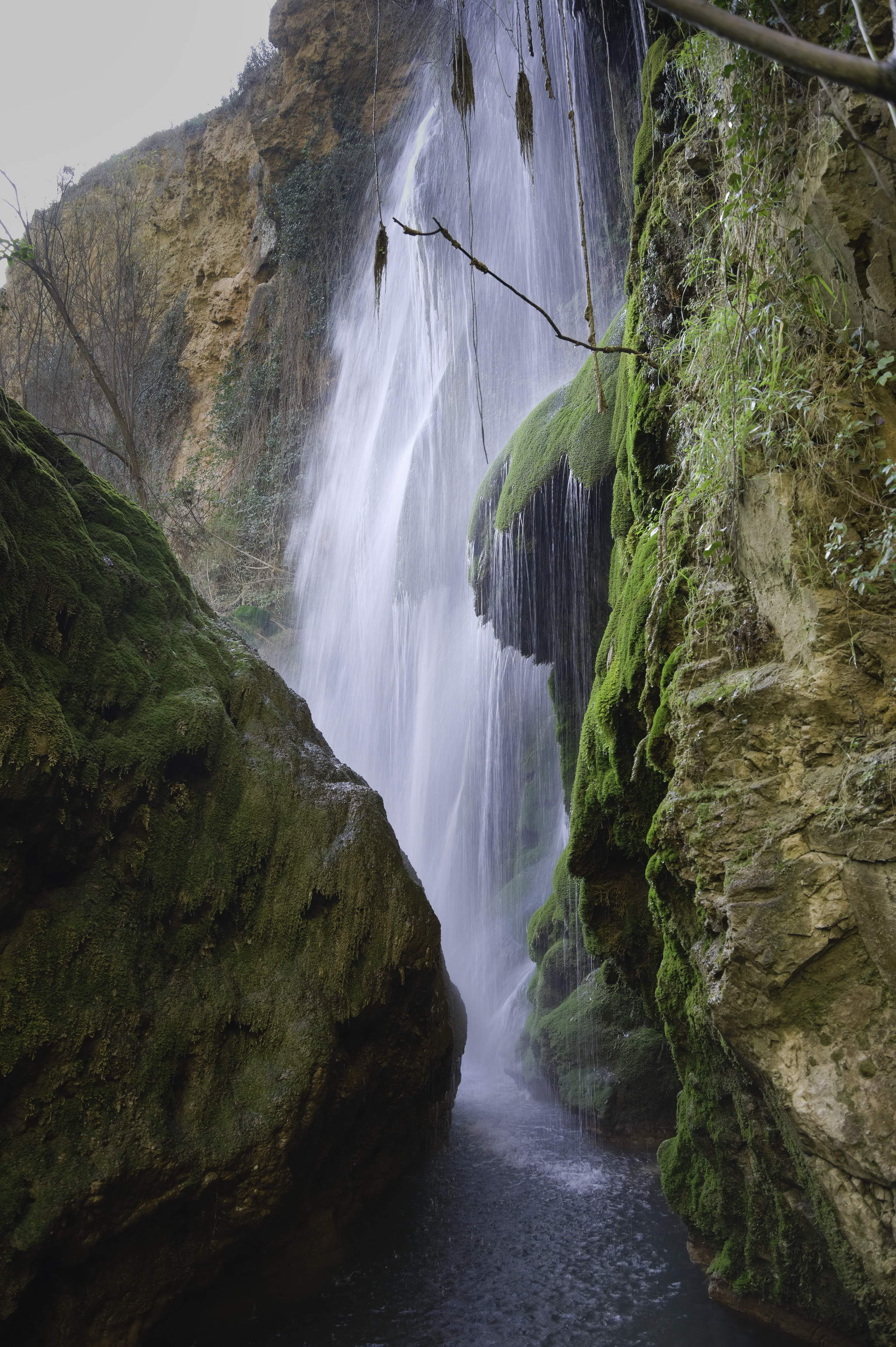 A huge waterfall drops down into a dark, green valley with lichen covered rocks.