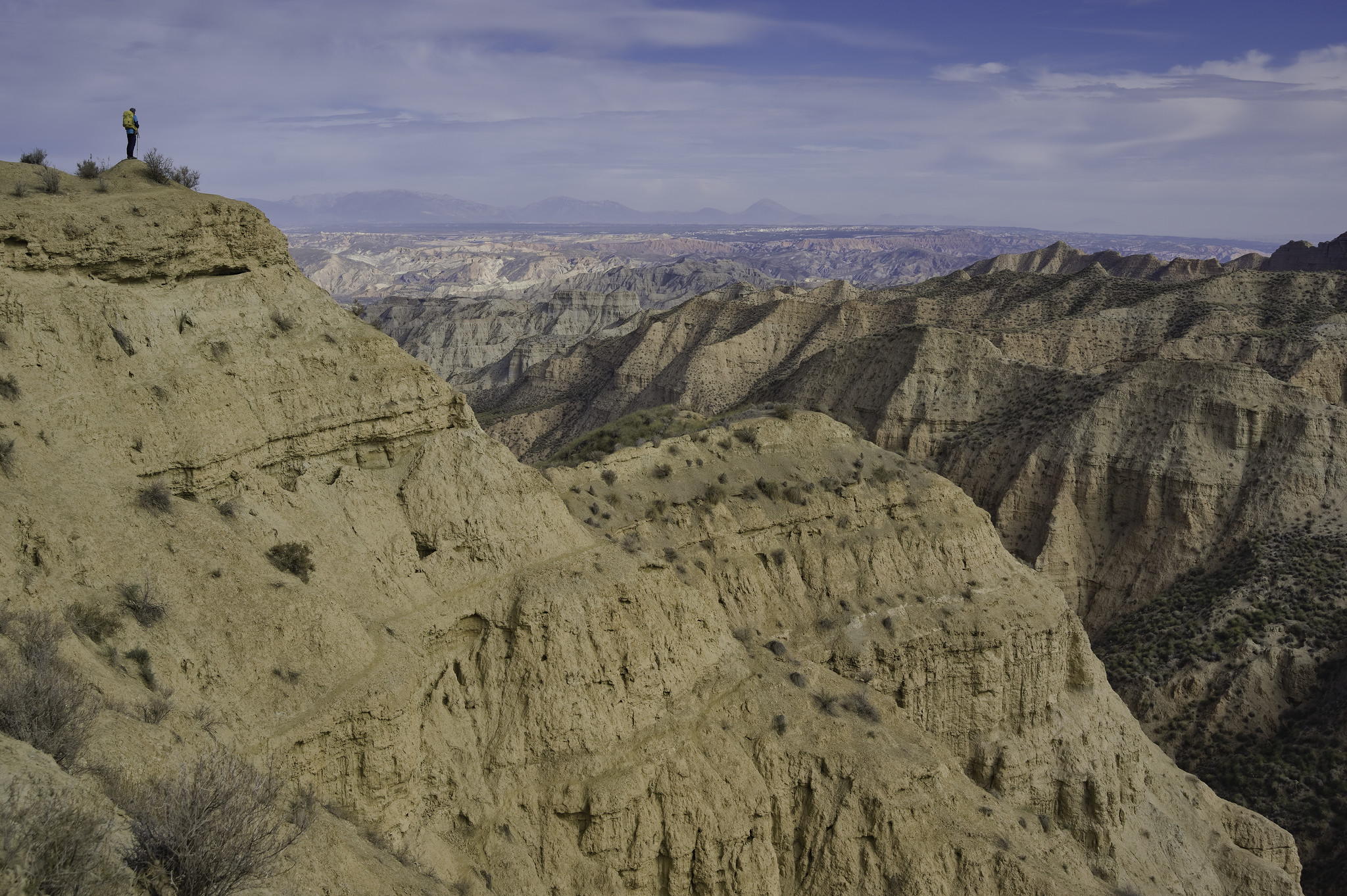 An isolated hiker stands in the top left of the photo. below and to the right is a huge drop into a twisted and tortured desert badlands terrain