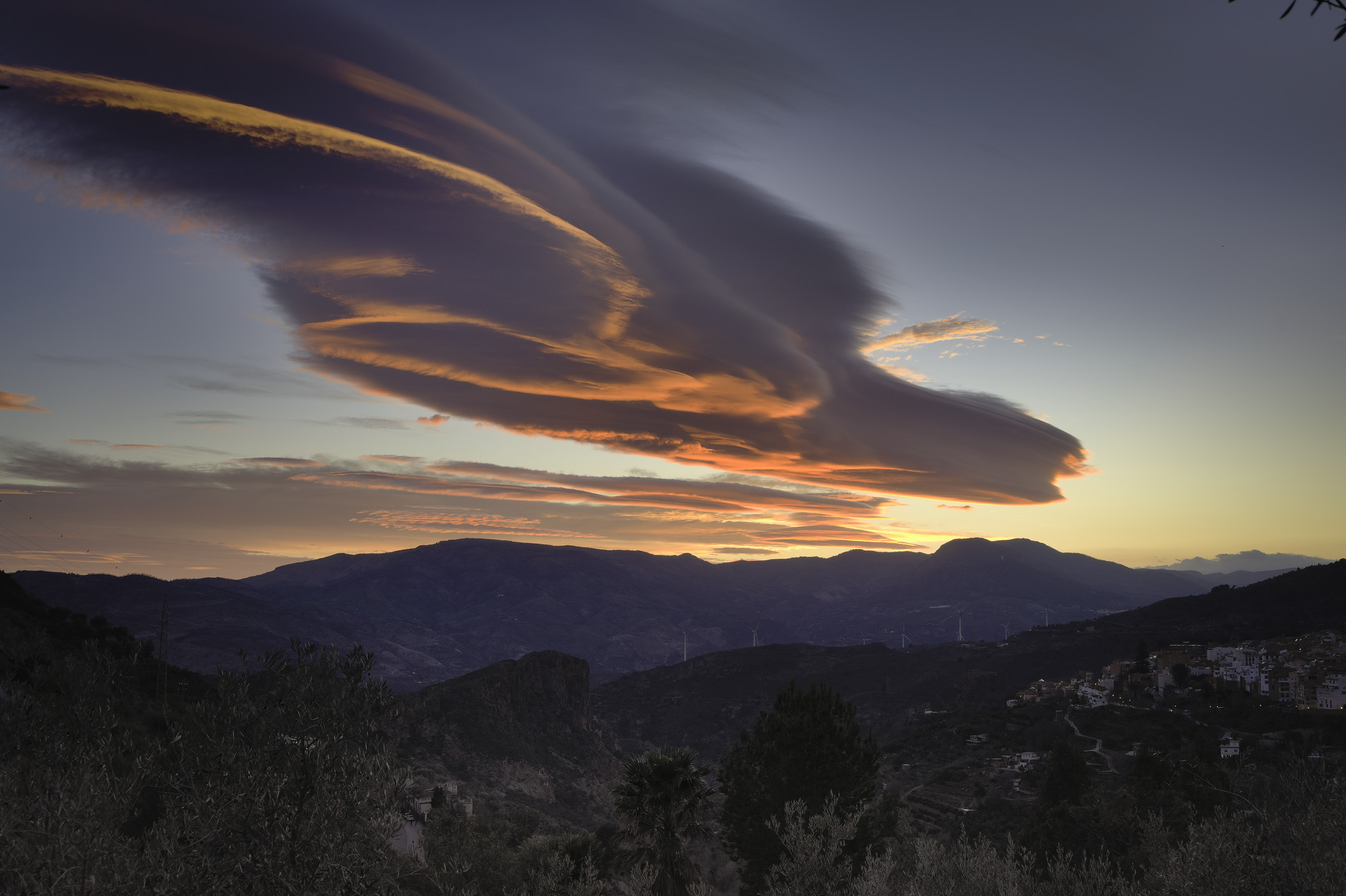 A huge orange/red coloured lens shaped cloud hovers above a small spanish town lying in the shade beneath it.