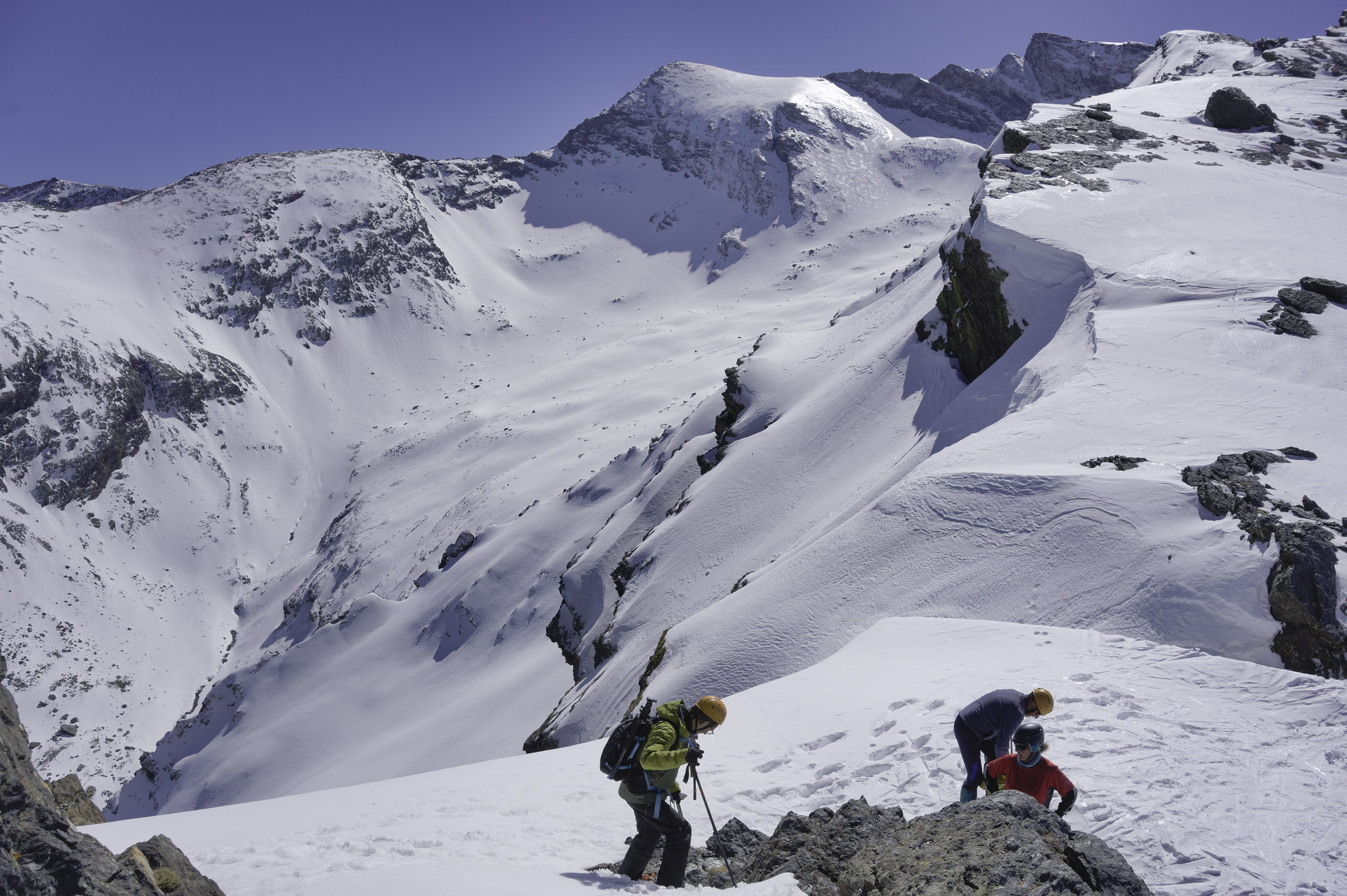 A group of mountaineering at the bottom of the image contemplate their way along the the snowy ridge ahead. Some big drops and high mountains to the left