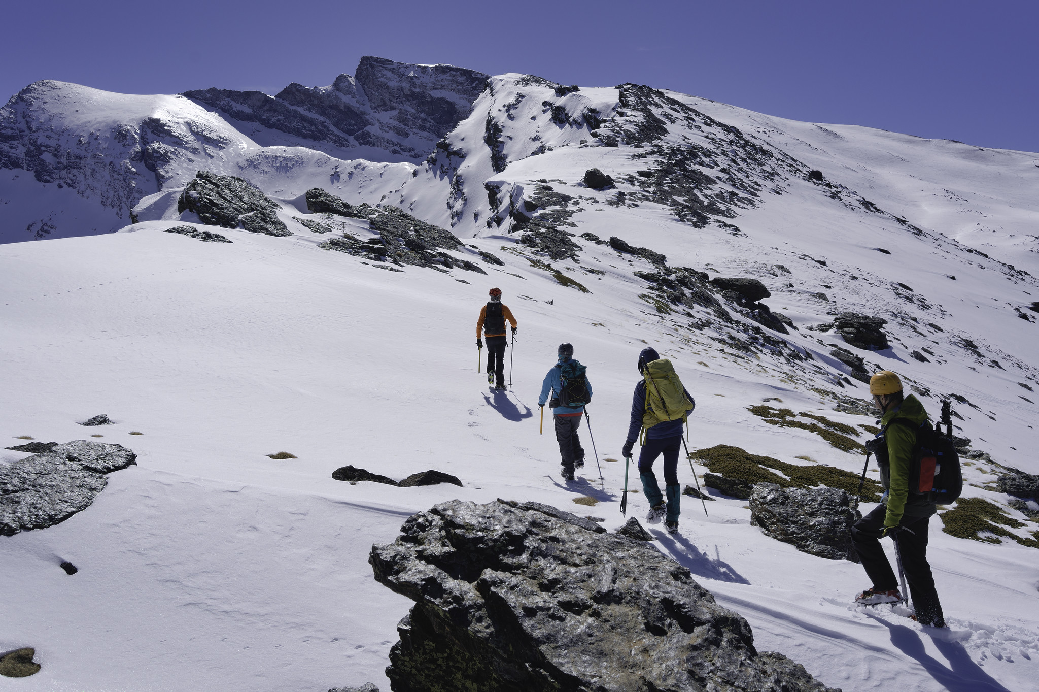 Four mountaineers dressed in colorful clothing make their way across some snow. Ahead lies a ridge leading to a jagged mountain