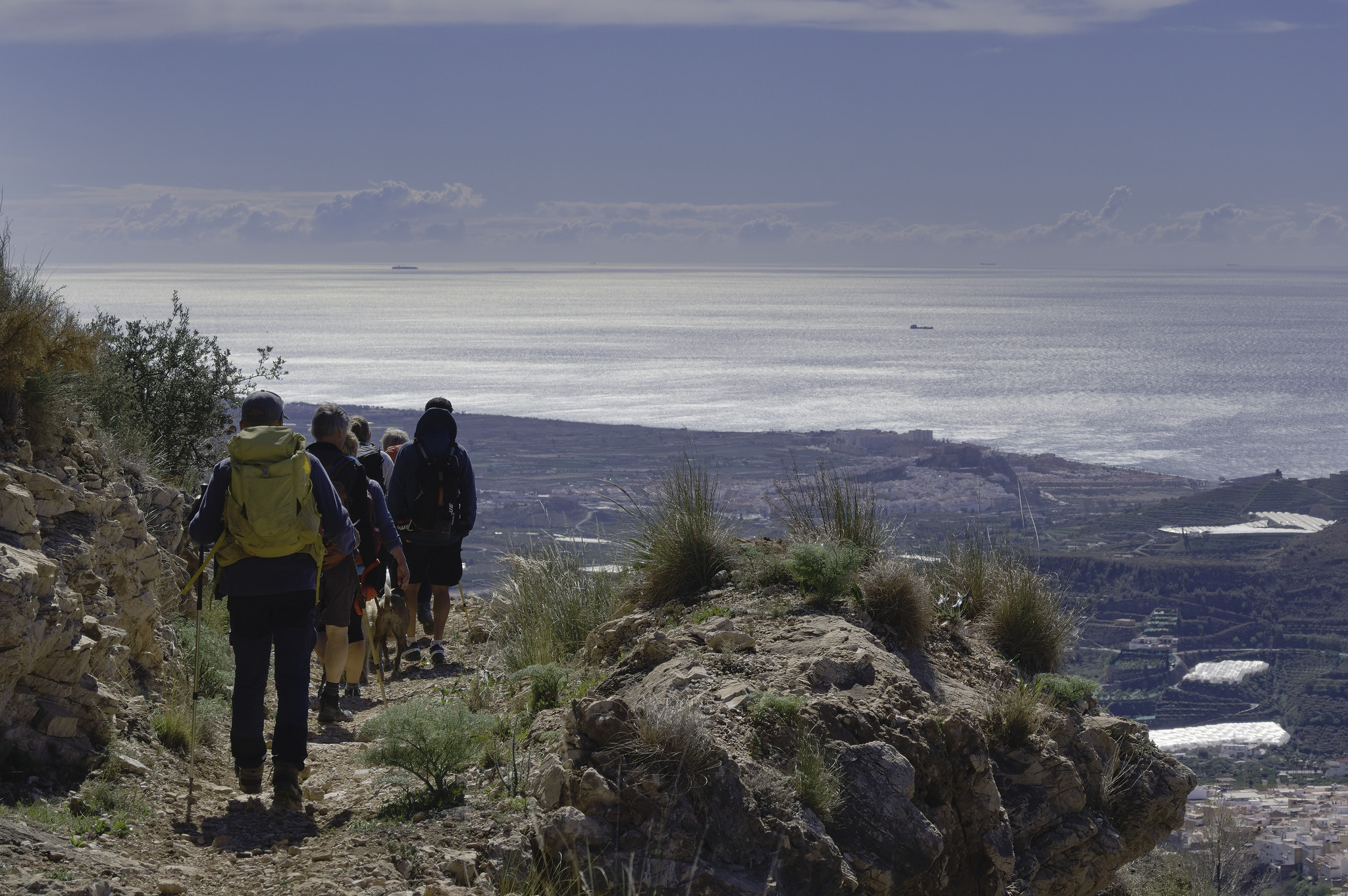 A group of hikers wander along a trail. In front of them lies the Mediterranean sea with a couple of ships on it.