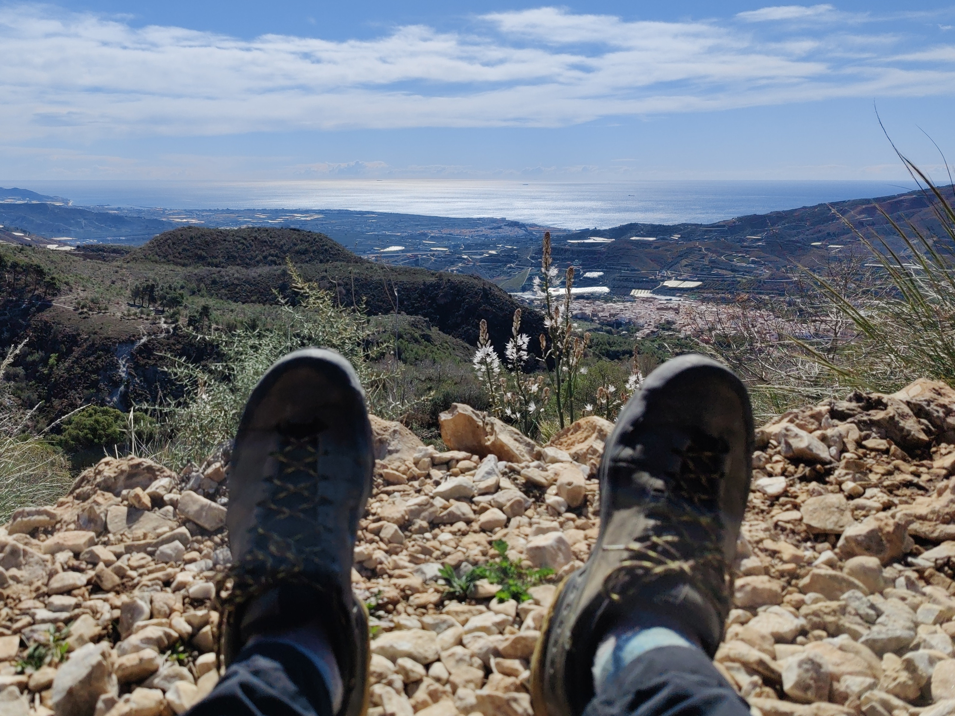 A pair of feet look out over an expansive view of green hills and towns. The Mediterranean Sea lies in the distance