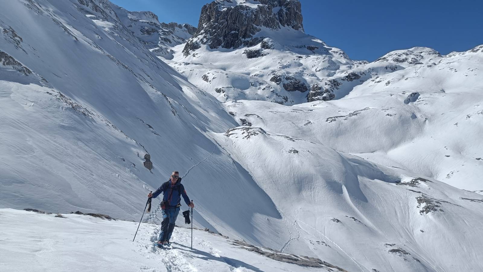 A person dressed in blue and in snowshoes is surrounded by snow and rugged high mountains. Blue sky above