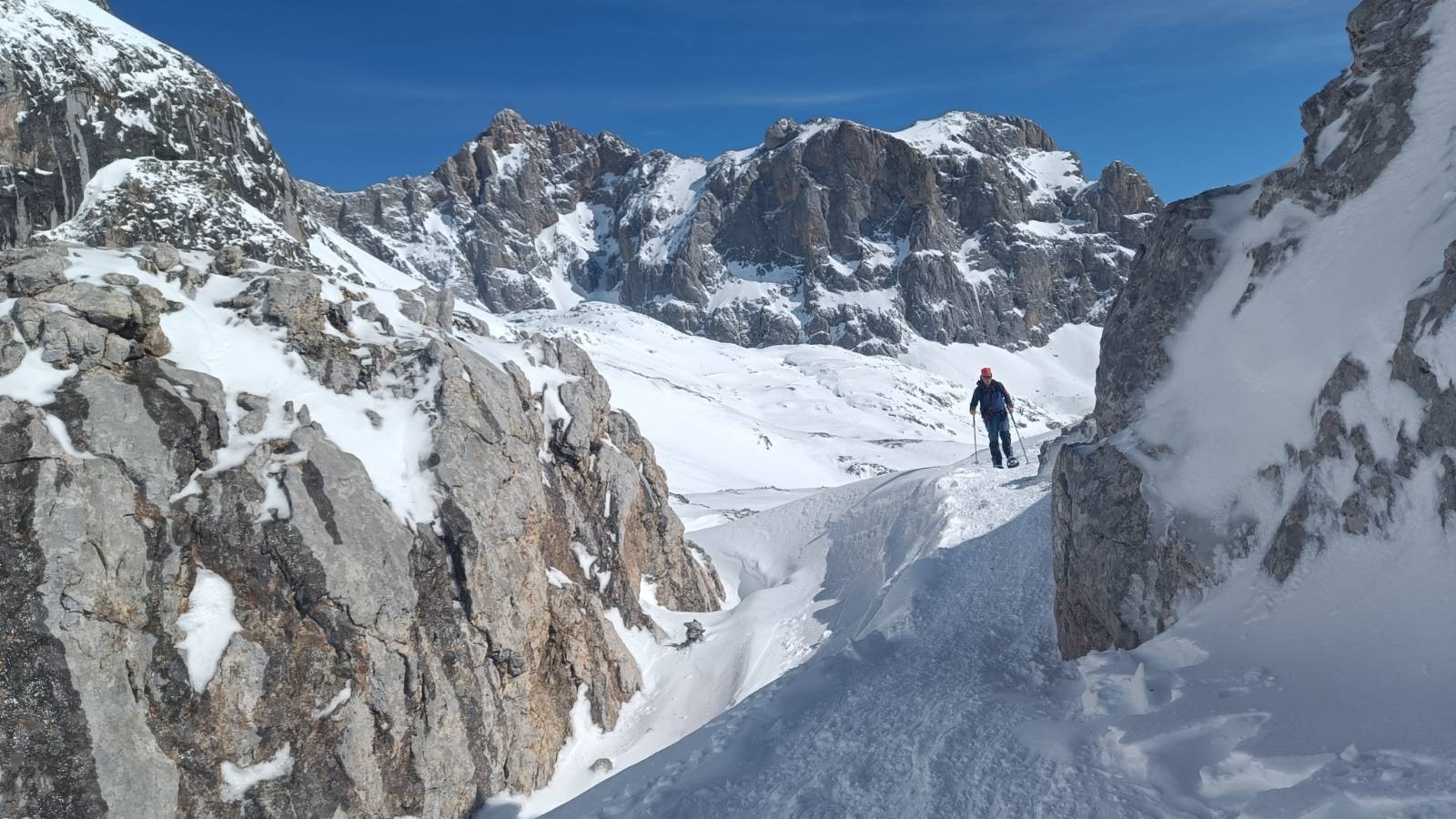 A person dressed in blue and in snowshoes is surrounded by snow and rugged high mountains. Blue sky above