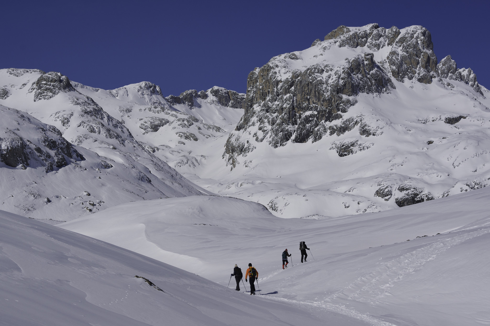 Some people with snowshoes and colorful clothing pass through undulating terrain heading for a distant valley surrounded by steep mountains