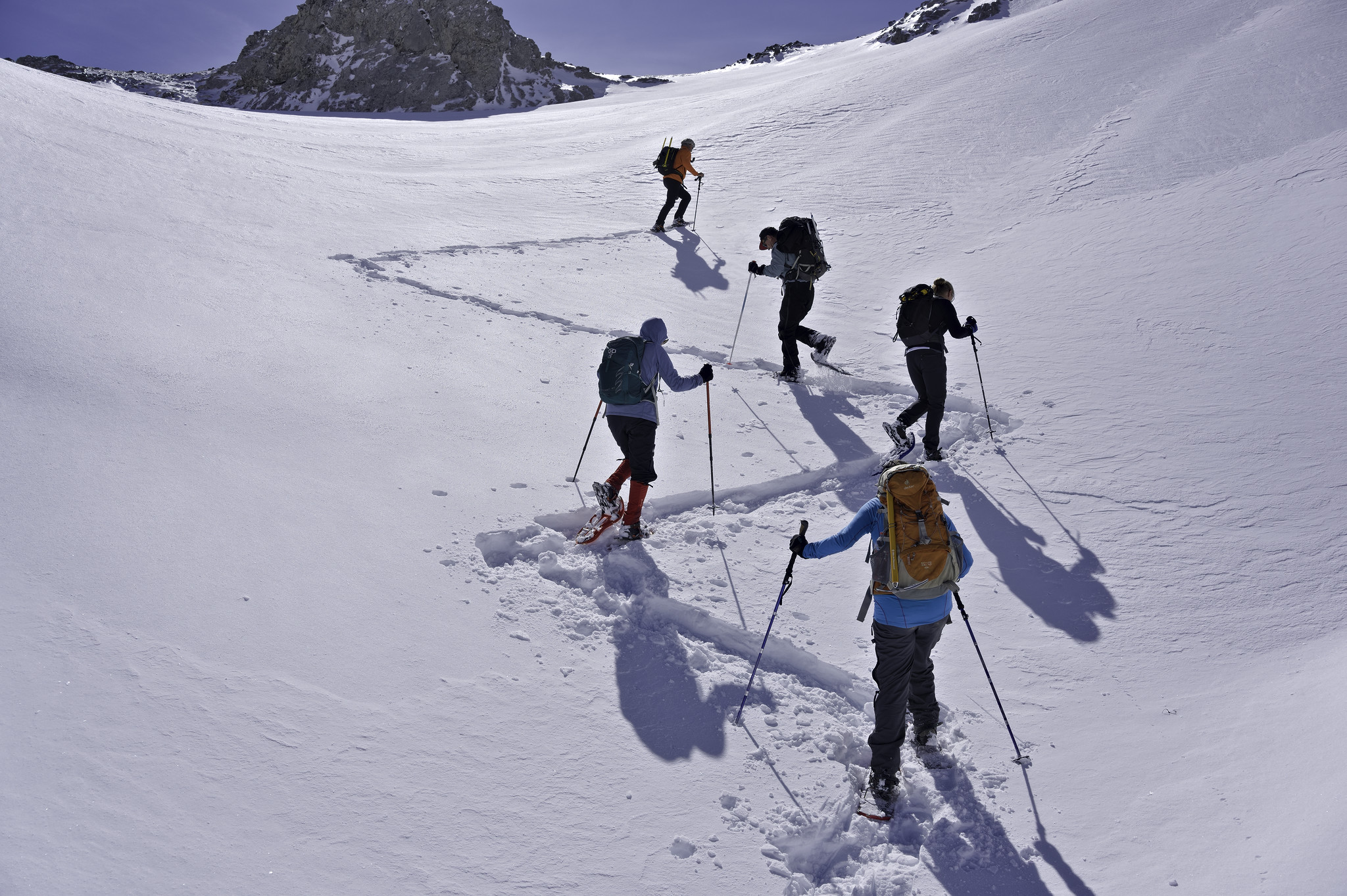5 people with snowshoes and colorful clothing zig zag up a steep snow slope