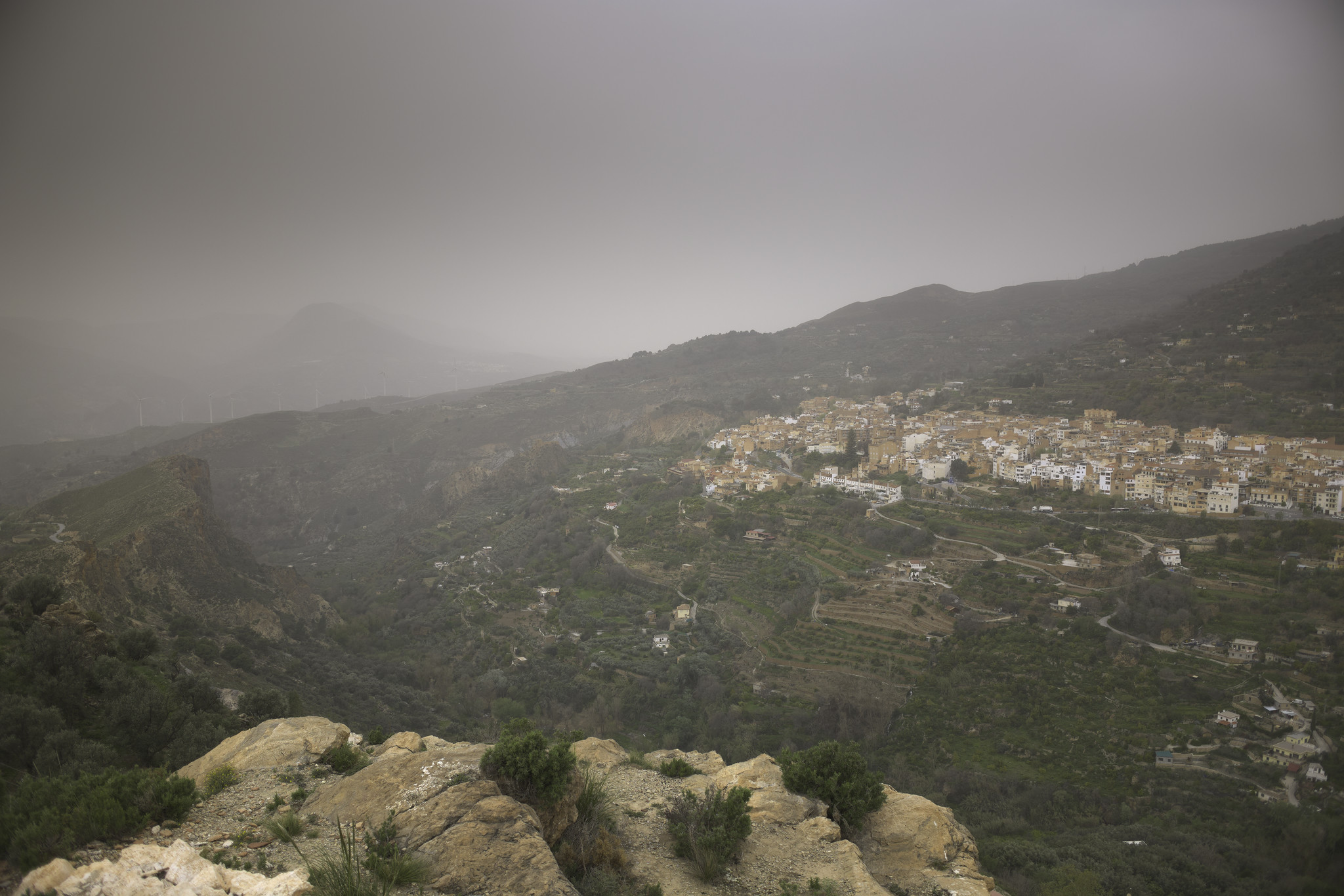 Out of the grey gloom a spanish town on a hill on the right hand side. To the left are green fields and some steep rocky crags