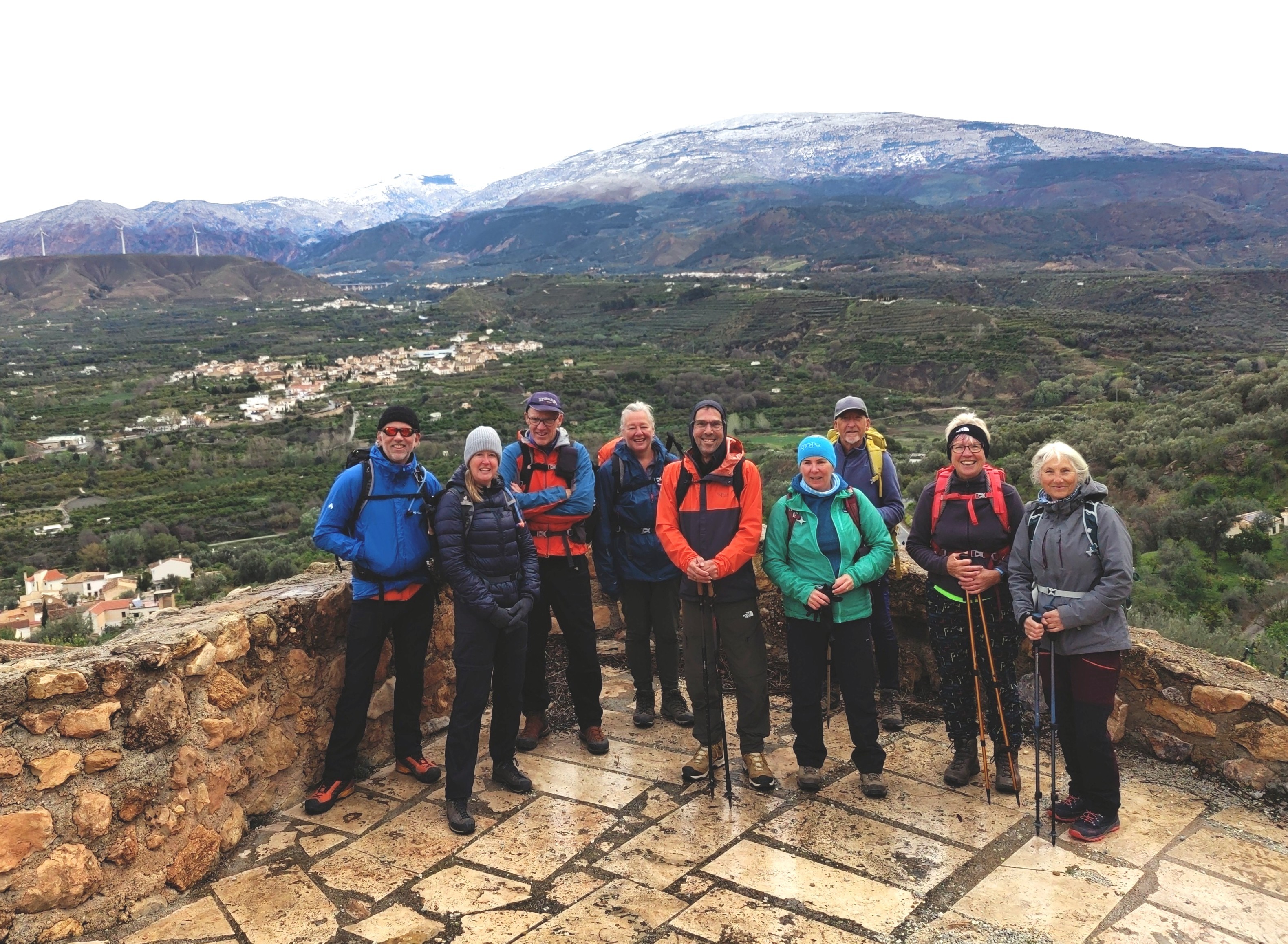 A group of hikers stand on a wet platform in damp conditions. Behind are villages and a mountainside blanketed by fresh snow