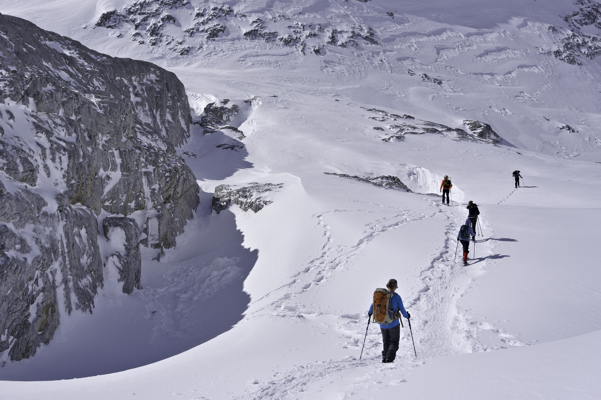 A group of 5 people snowshoeing int eh white snows are passing to the right of a shadowed icy cliff