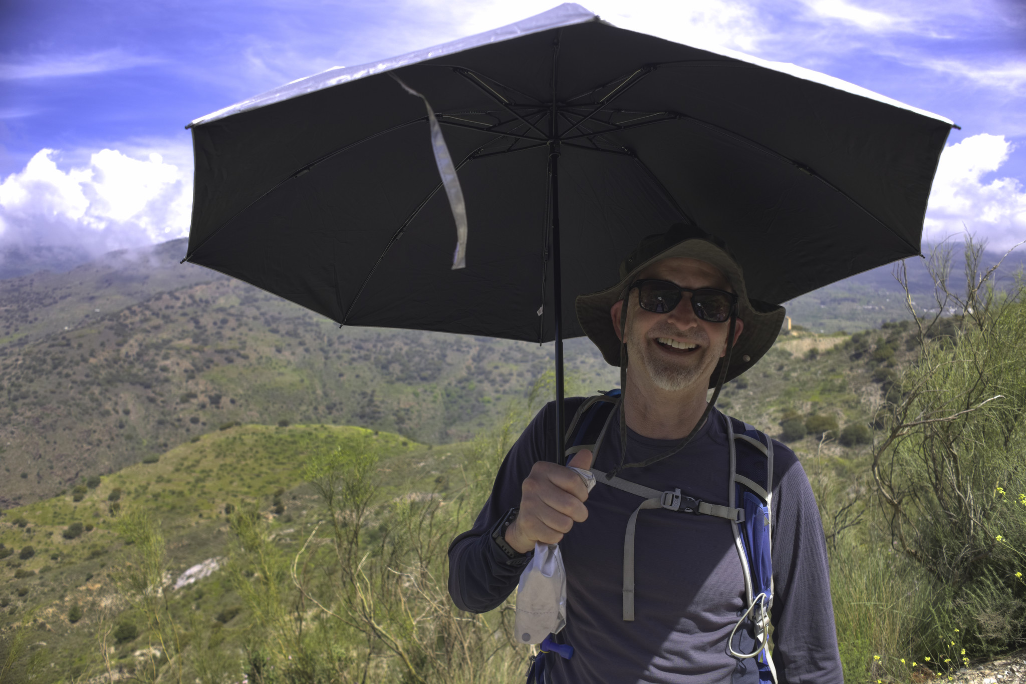 A smiling man stands under a black shaded umbrella. Above are blue skies and white clouds