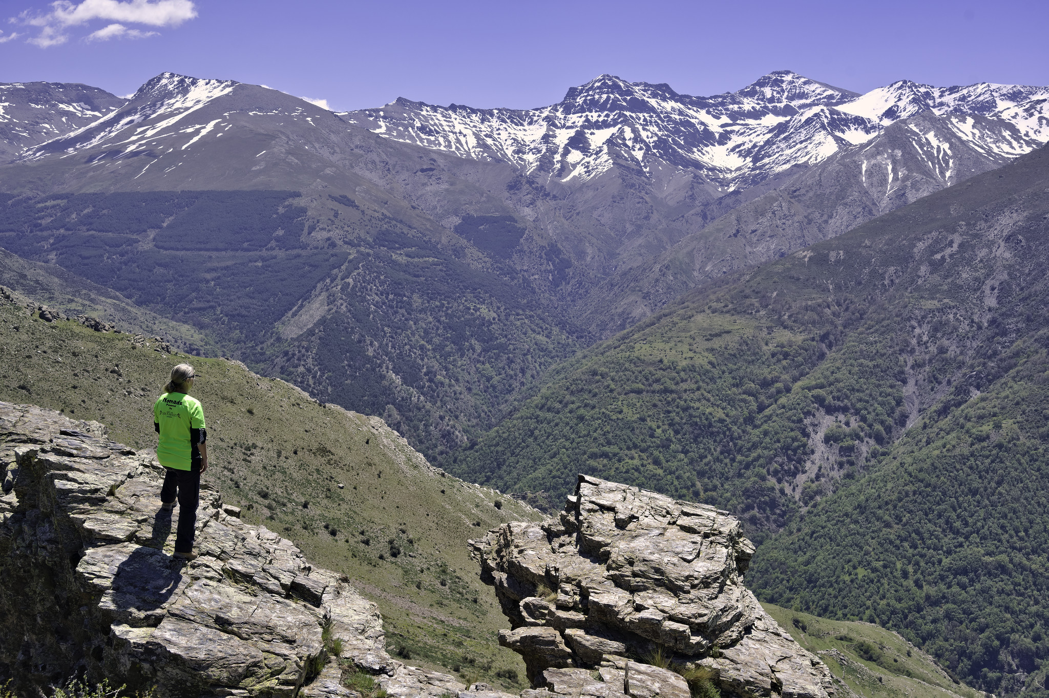A person is stood on a rock platform in the bottom left overlooking a deep valley with snowy mountains in the distance