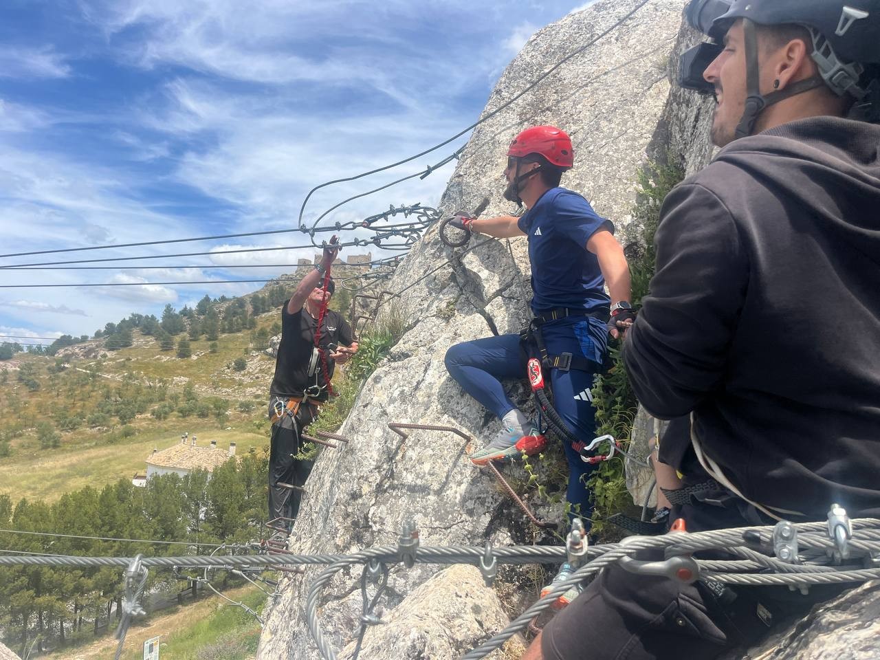 One person is securing himself to cables whilst one person with a red helmet watches. On the right of the image is a black dressed cameraman filming the scene