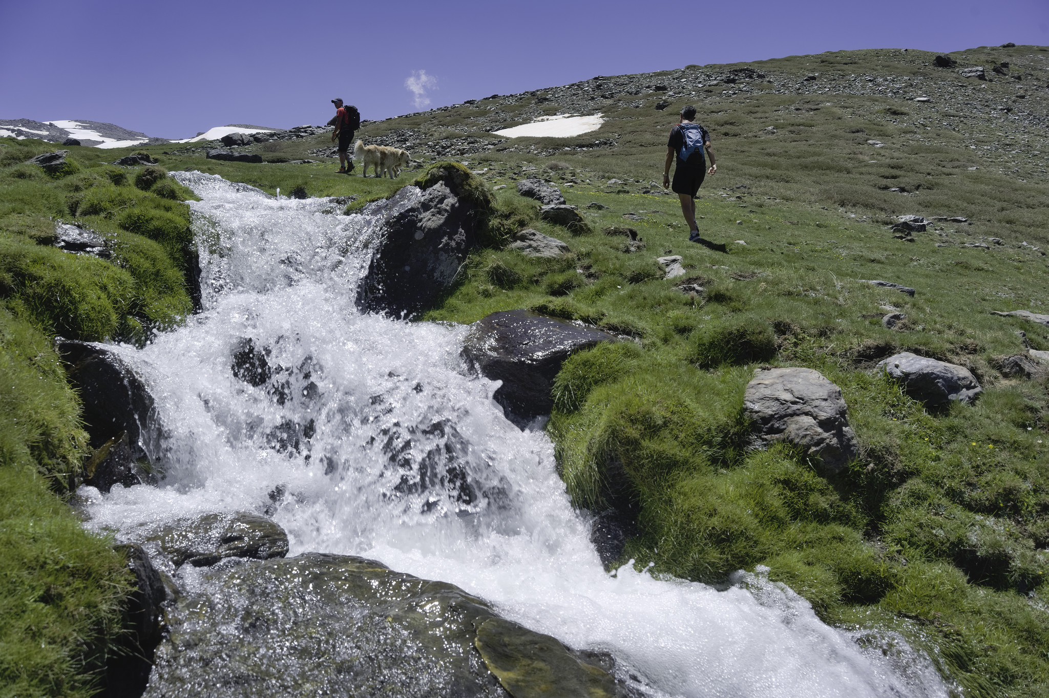A large stream gushes out of a lip in the valley. Two hikers are seen walking up the far grassy banks. There is a hint of higher snowy mountains above