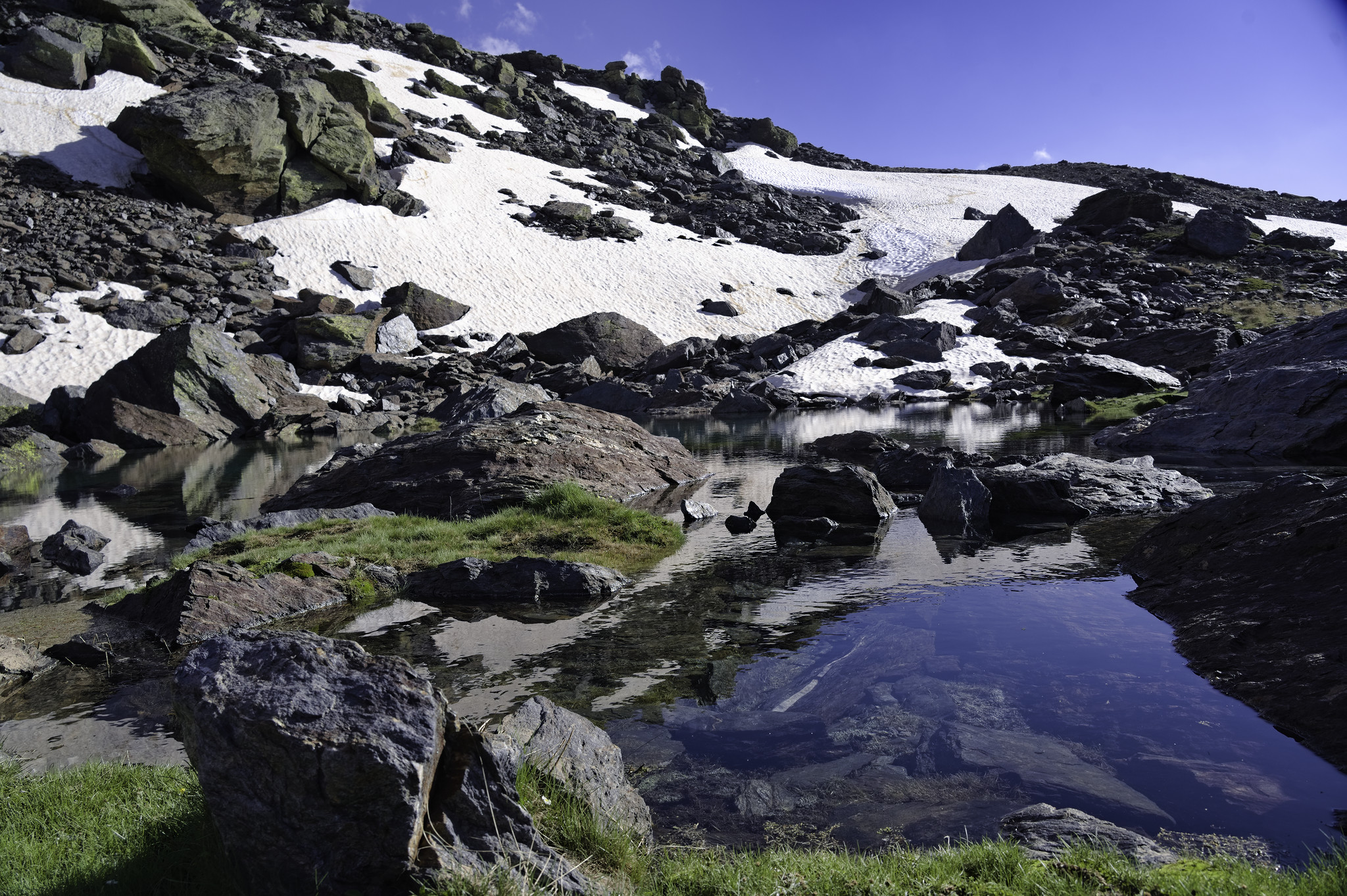 A lake with green banks is surrounded by cliffs and snow slopes. Blue sky above