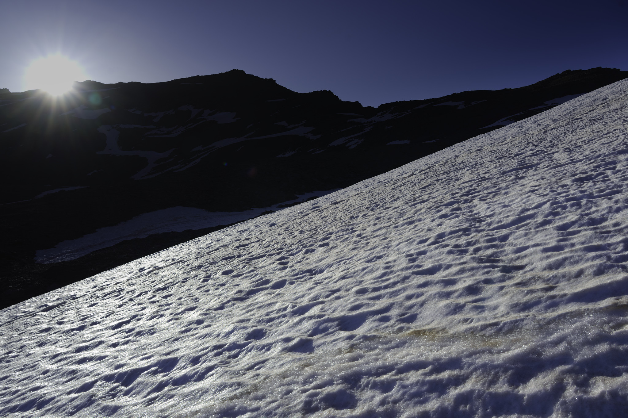 Out of the Shadows and into the Sun. A snow slope is highlighted by the rising sun coming over a black mountain ridge
