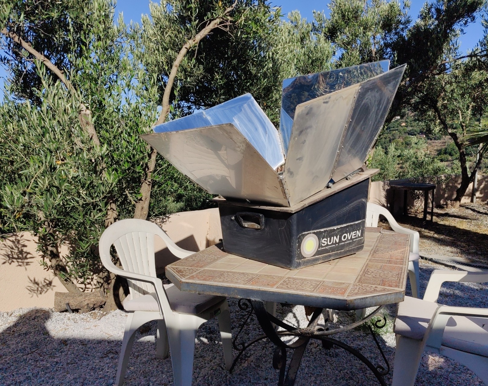 A solar oven with a parabolic shield sits on a table surrounded by chairs. In the background some olive trees.